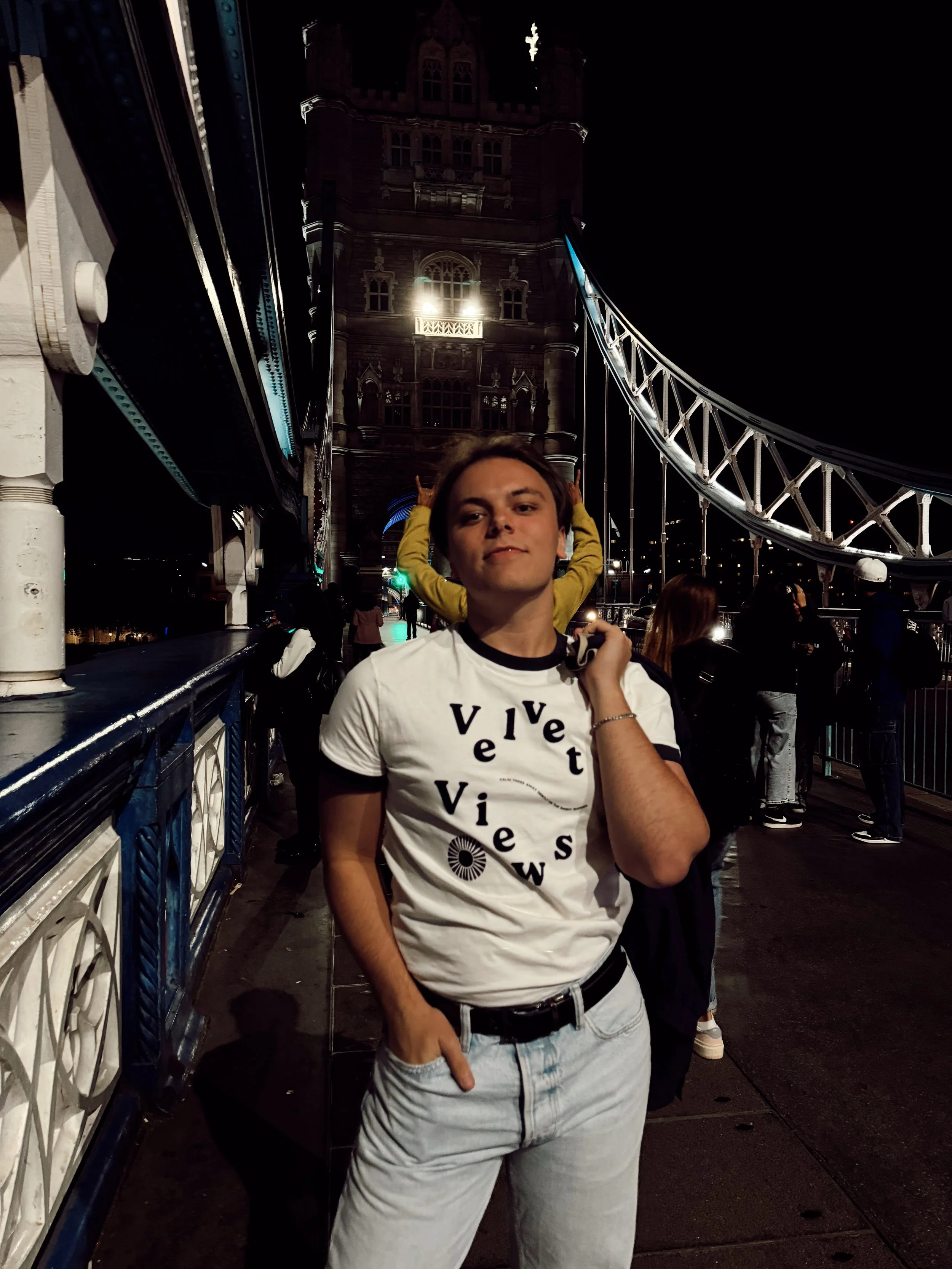 A young man in a white t-shirt with black text and light-colored jeans stands on a bridge at night, with Tower Bridge in London illuminated in the background.