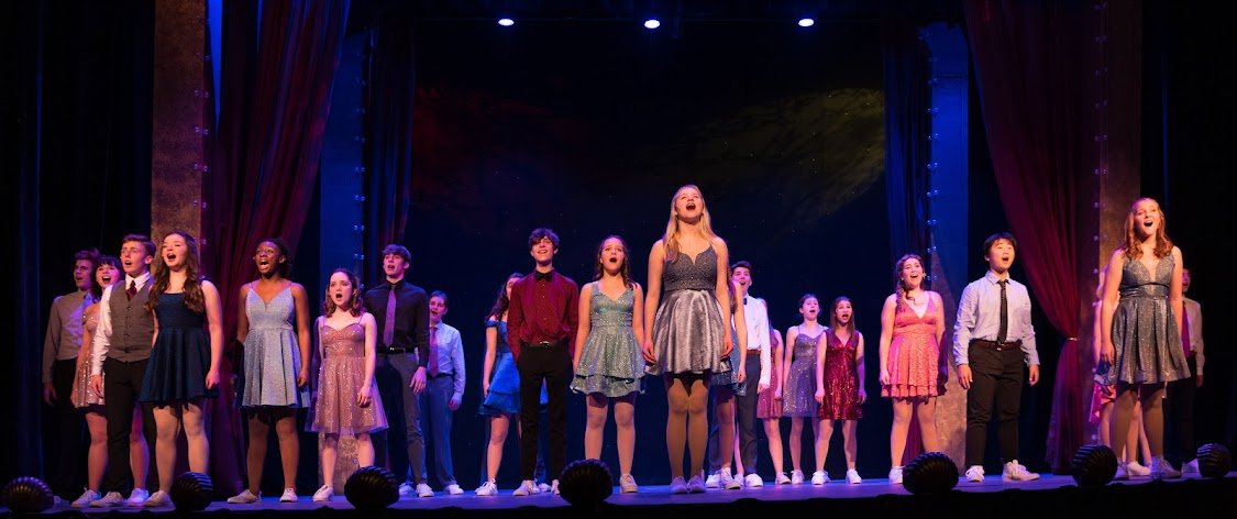 A group of young performers on stage singing during a theatrical production, with some holding hands and others with hands at their sides, in front of red curtains and colorful stage lighting.