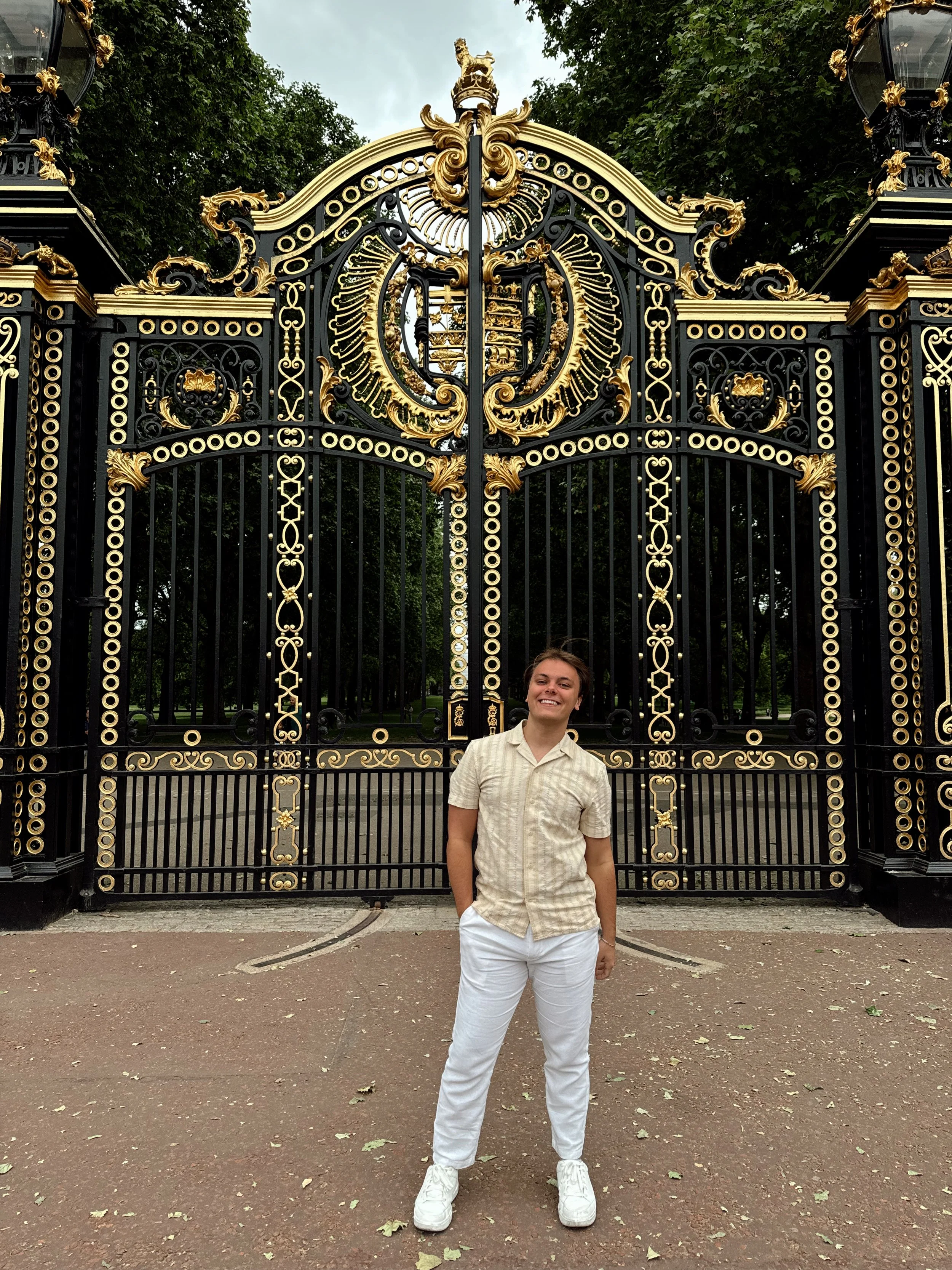 A young man standing in front of an ornate black and gold wrought-iron gate, smiling.