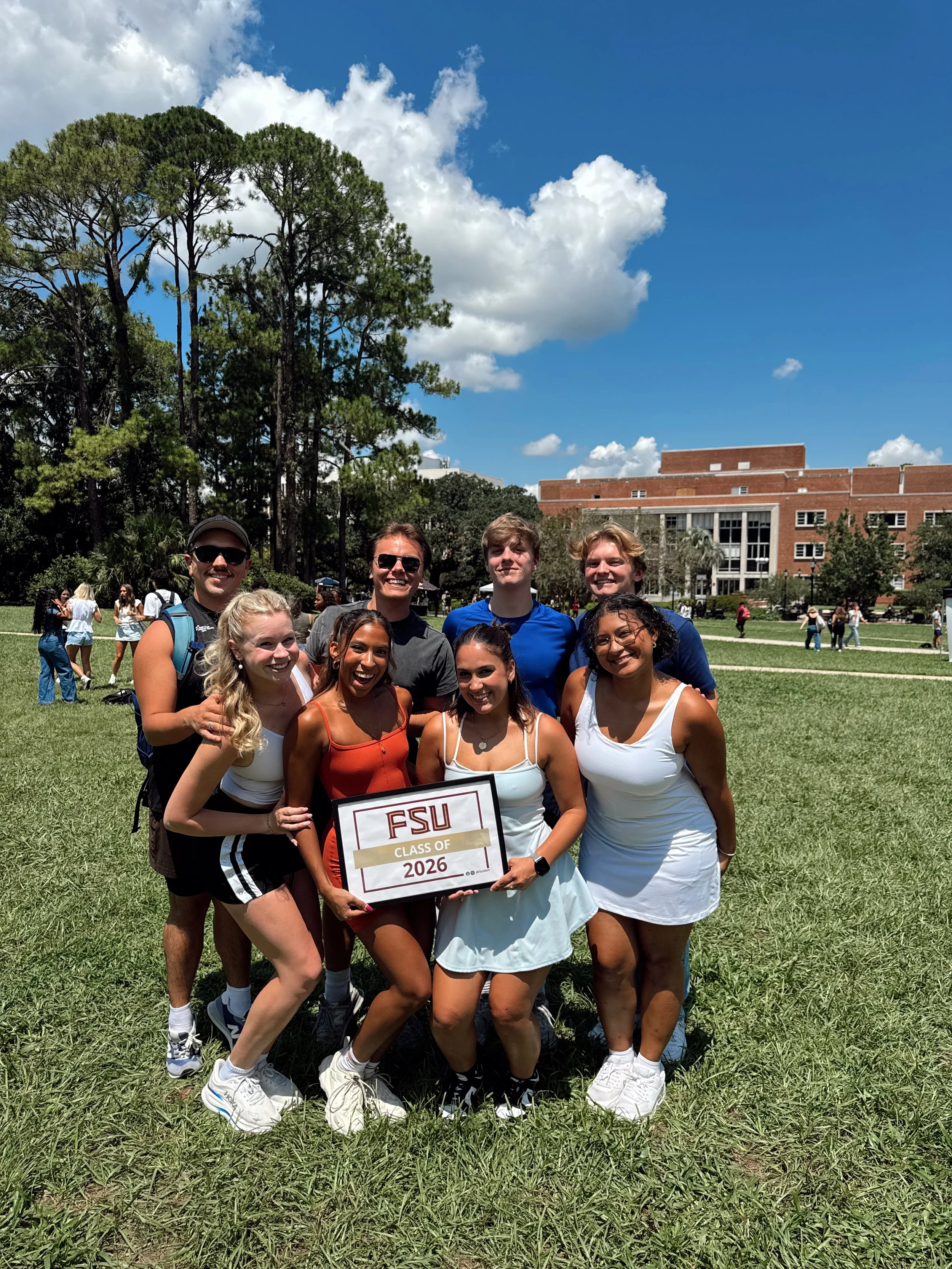 Group of eight diverse young adults, smiling, holding a sign that reads "FSU Class of 2026" on a college campus grassy field with trees and a large brick building in the background on a sunny day.