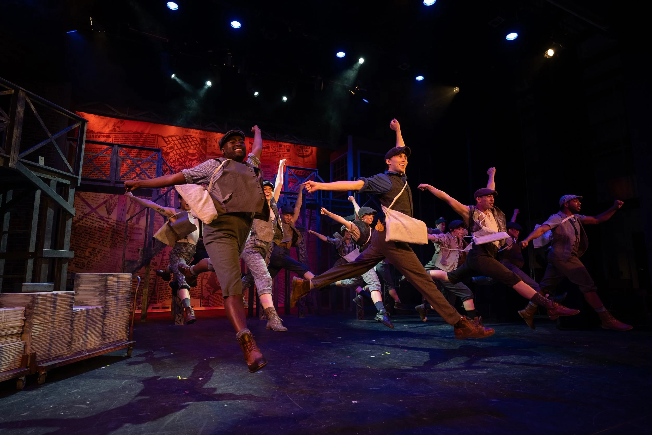 Theater performers acting in a scene with joyful expressions, dressed in vintage work attire, in a stage setting with industrial props and a backdrop resembling old newspapers.