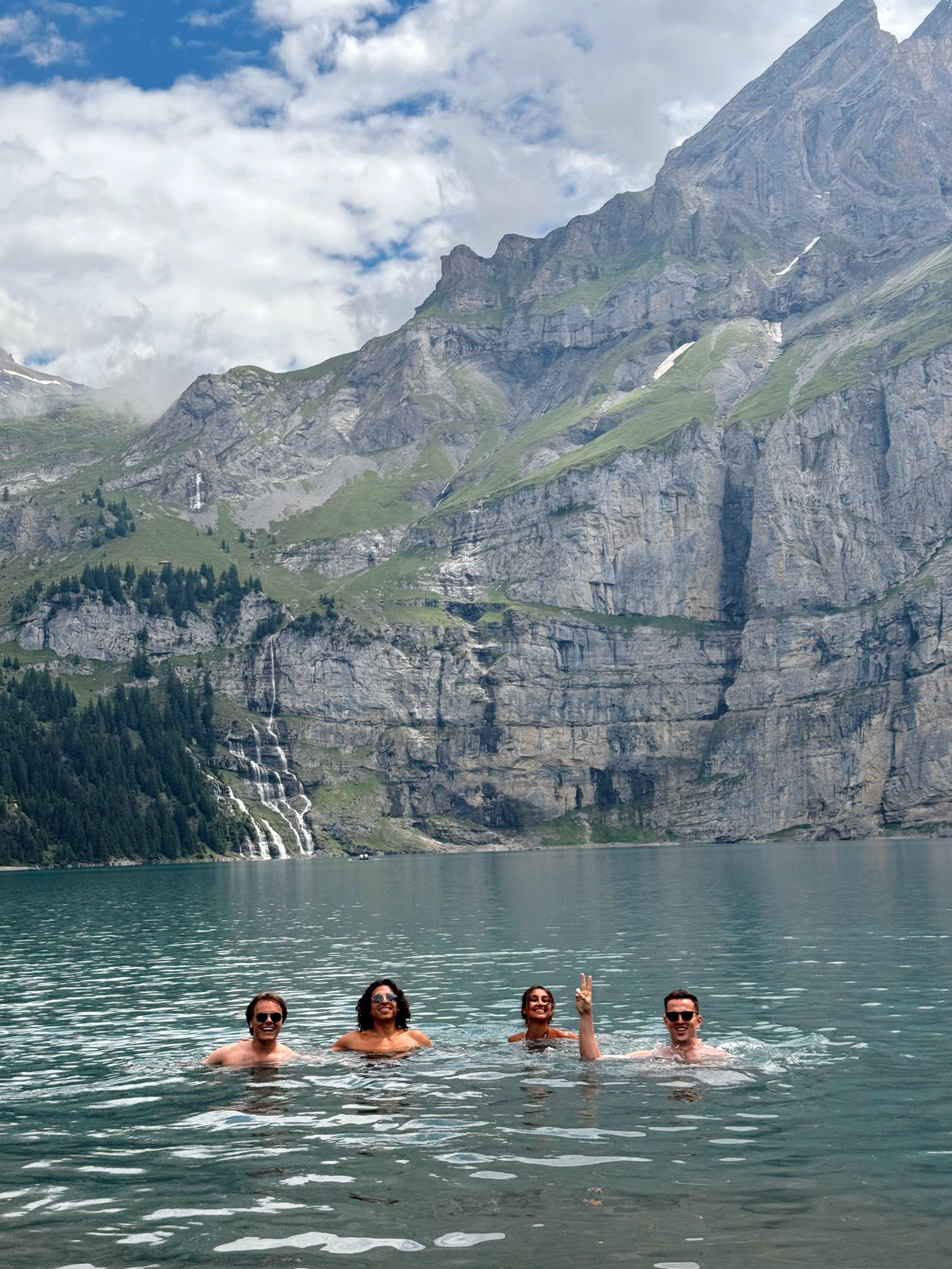 Four people swimming in a lake with mountain cliffs and waterfalls in the background.