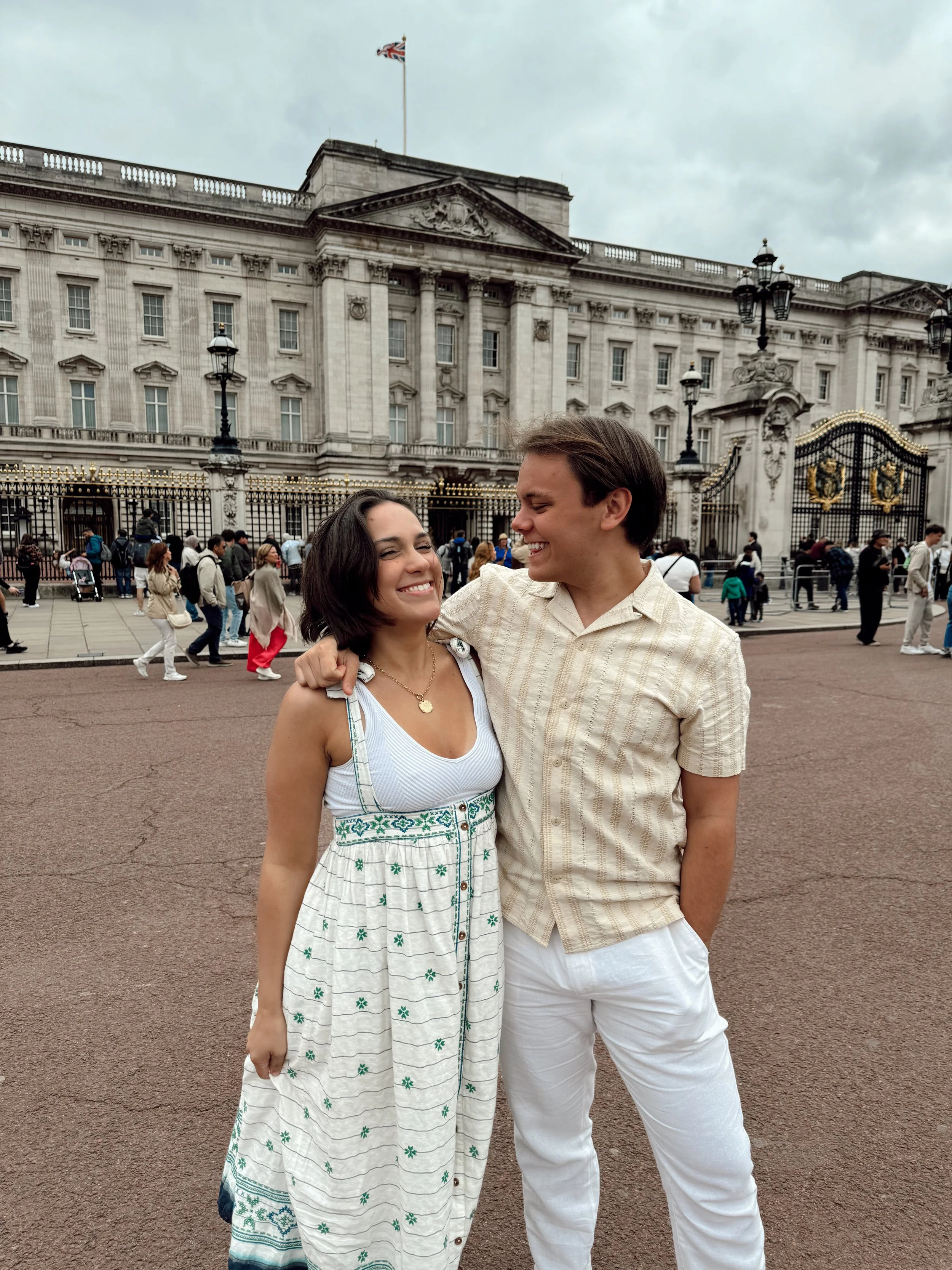 A young couple smiling and embracing in front of Buckingham Palace in London, England, with tourists in the background and the Union Jack flag flying above the palace.