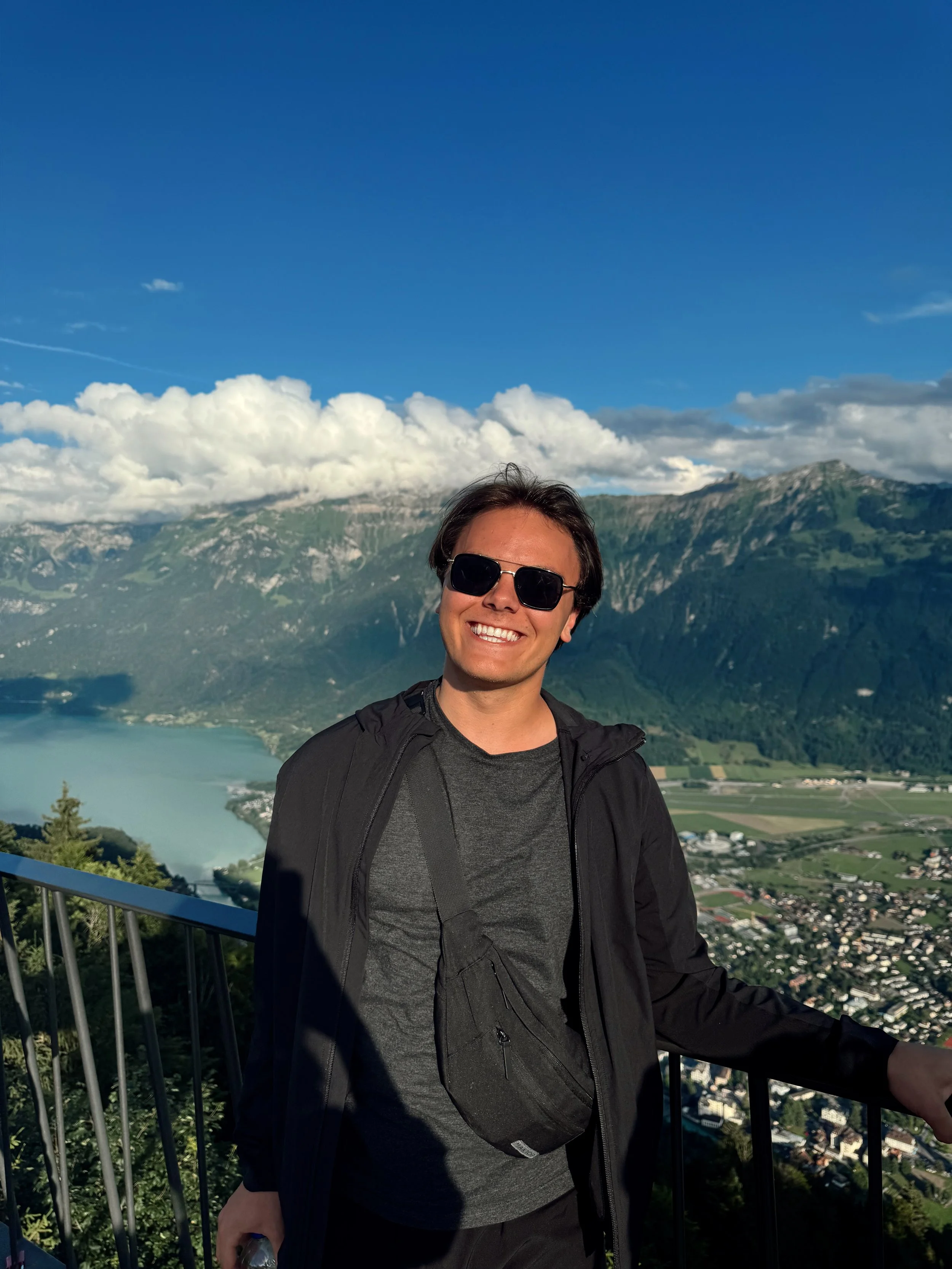 Smiling man wearing sunglasses and a black jacket standing on a balcony with a scenic mountainous landscape and lake in the background.