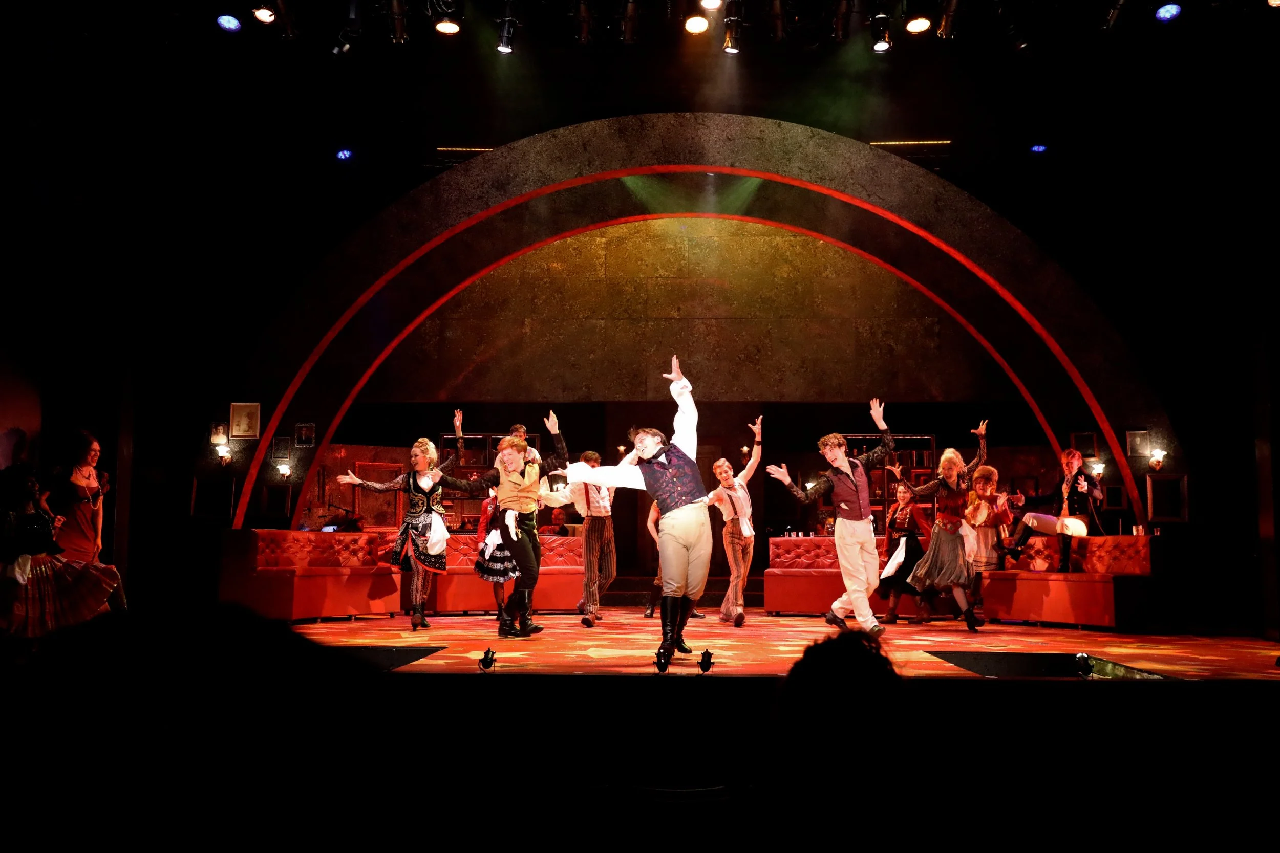 Stage scene from a theatrical performance with actors dancing and singing, illuminated by stage lights with a decorated background.