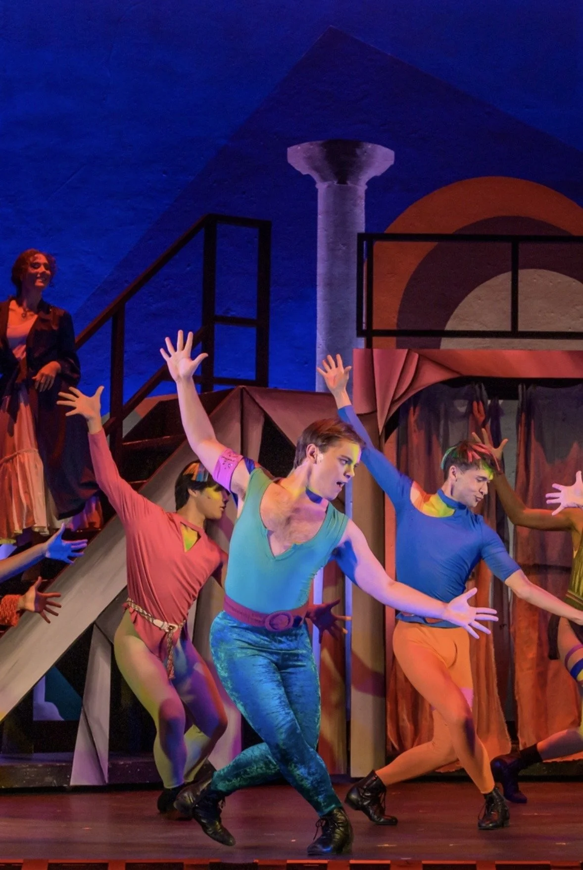 Performers dancing on stage in colorful costumes with a geometric set design and blue lighting.