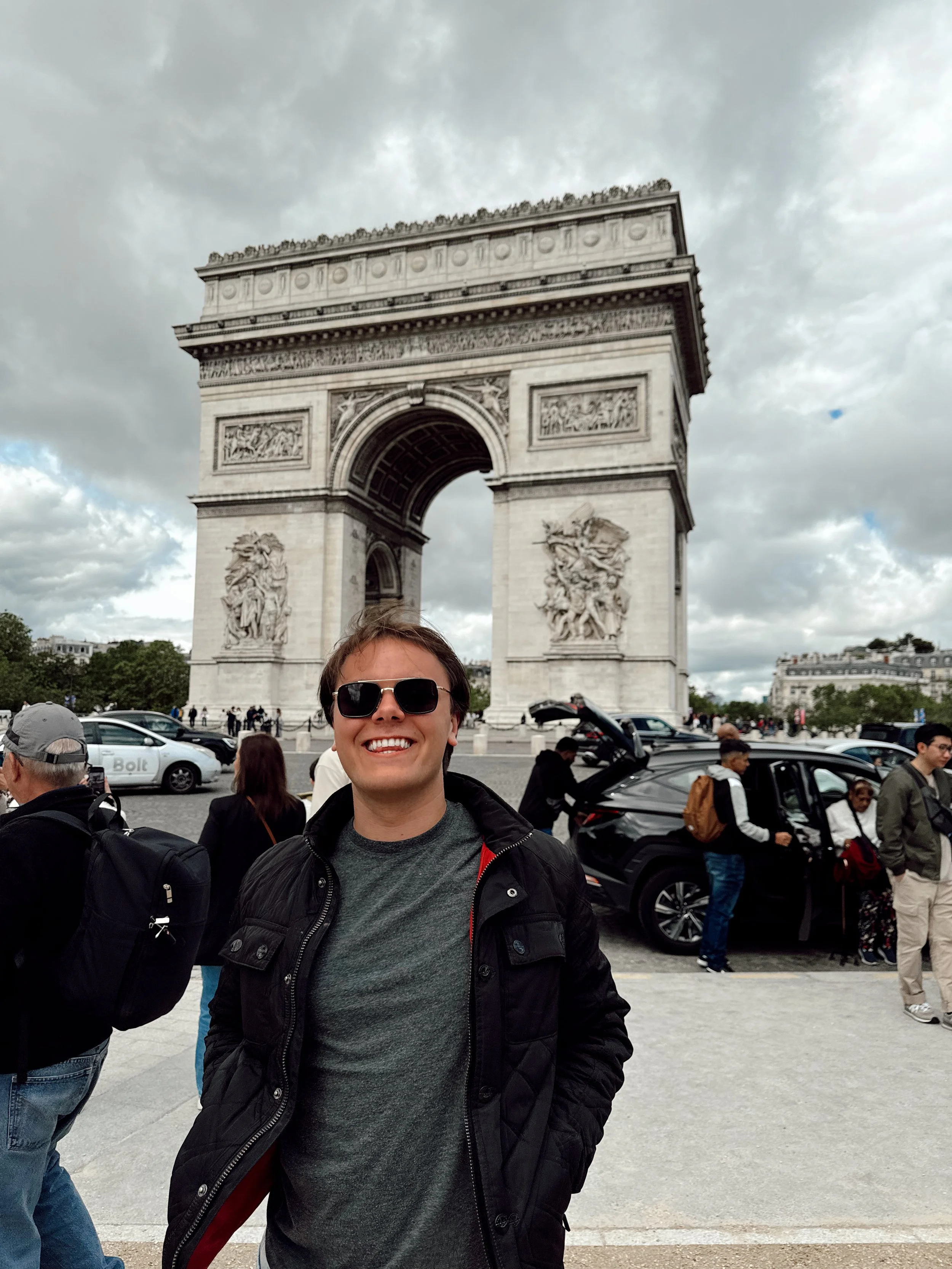 A smiling man wearing sunglasses and a black jacket posing in front of the Arc de Triomphe in Paris, with people and cars nearby on a cloudy day.