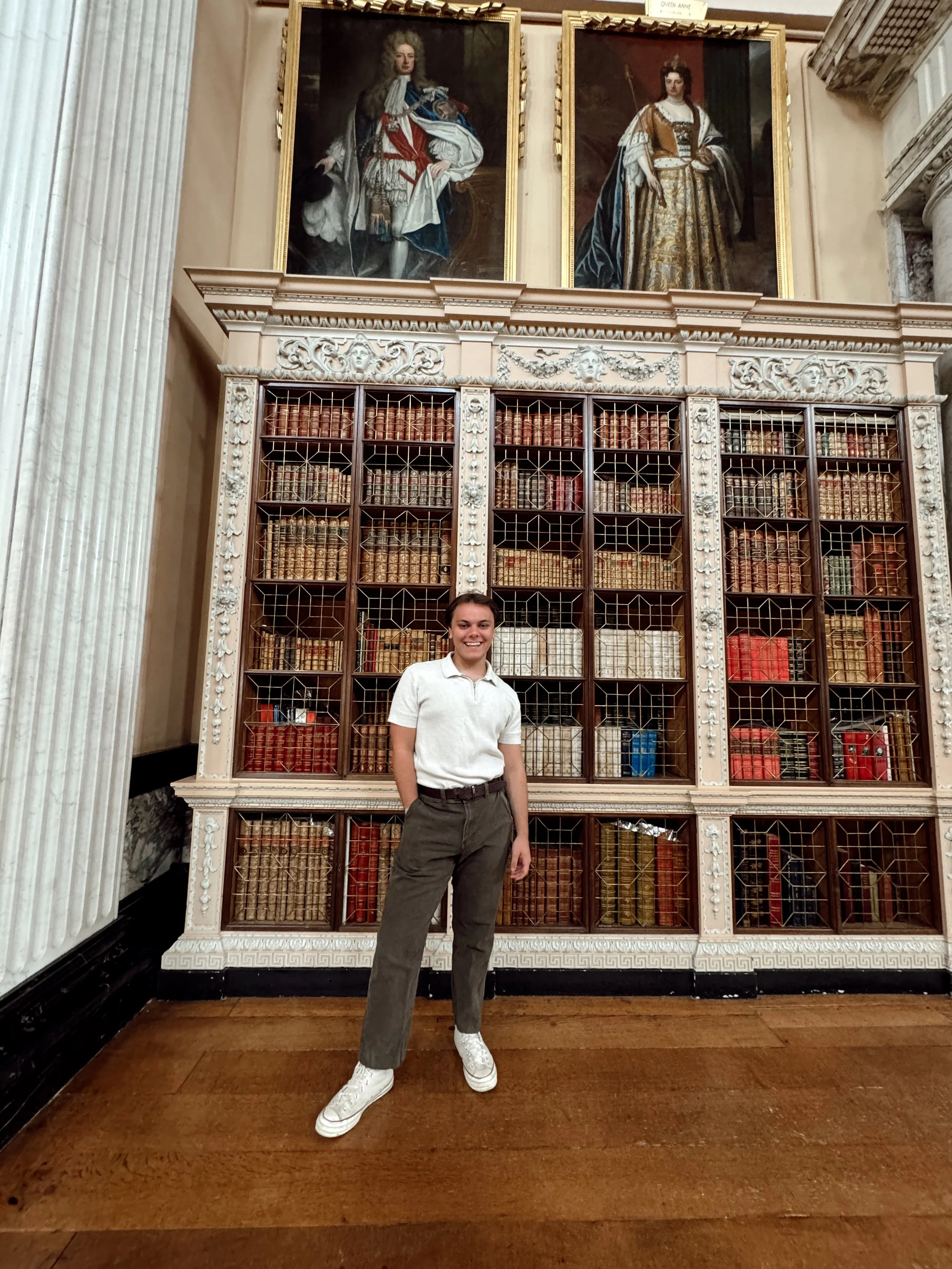 A woman standing in front of a large ornate bookcase filled with old books, in a historic library or museum with two large portrait paintings hanging above the bookcase.