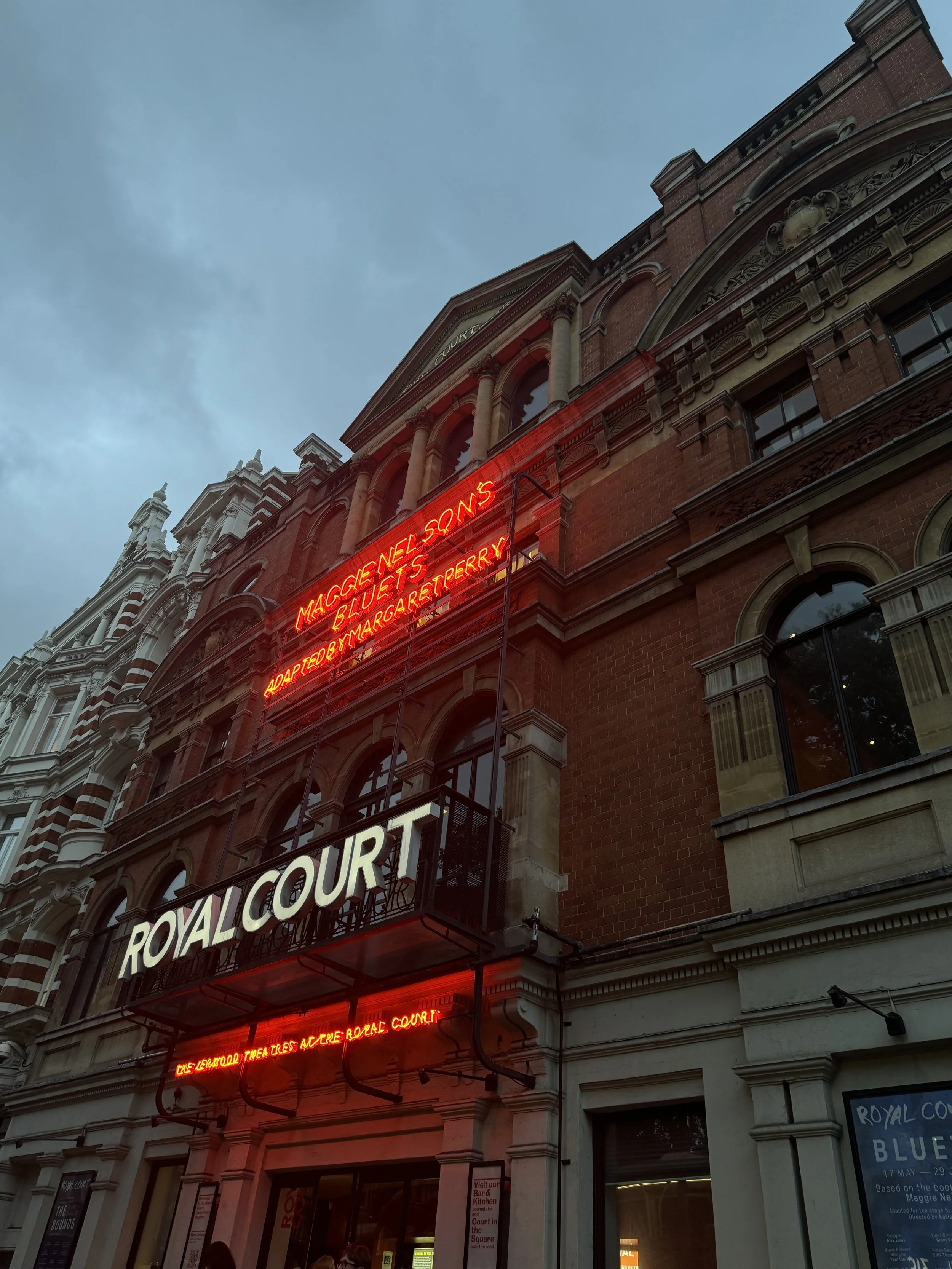 Exterior view of the Royal Court building with illuminated red neon signs displaying show titles, under cloudy sky.