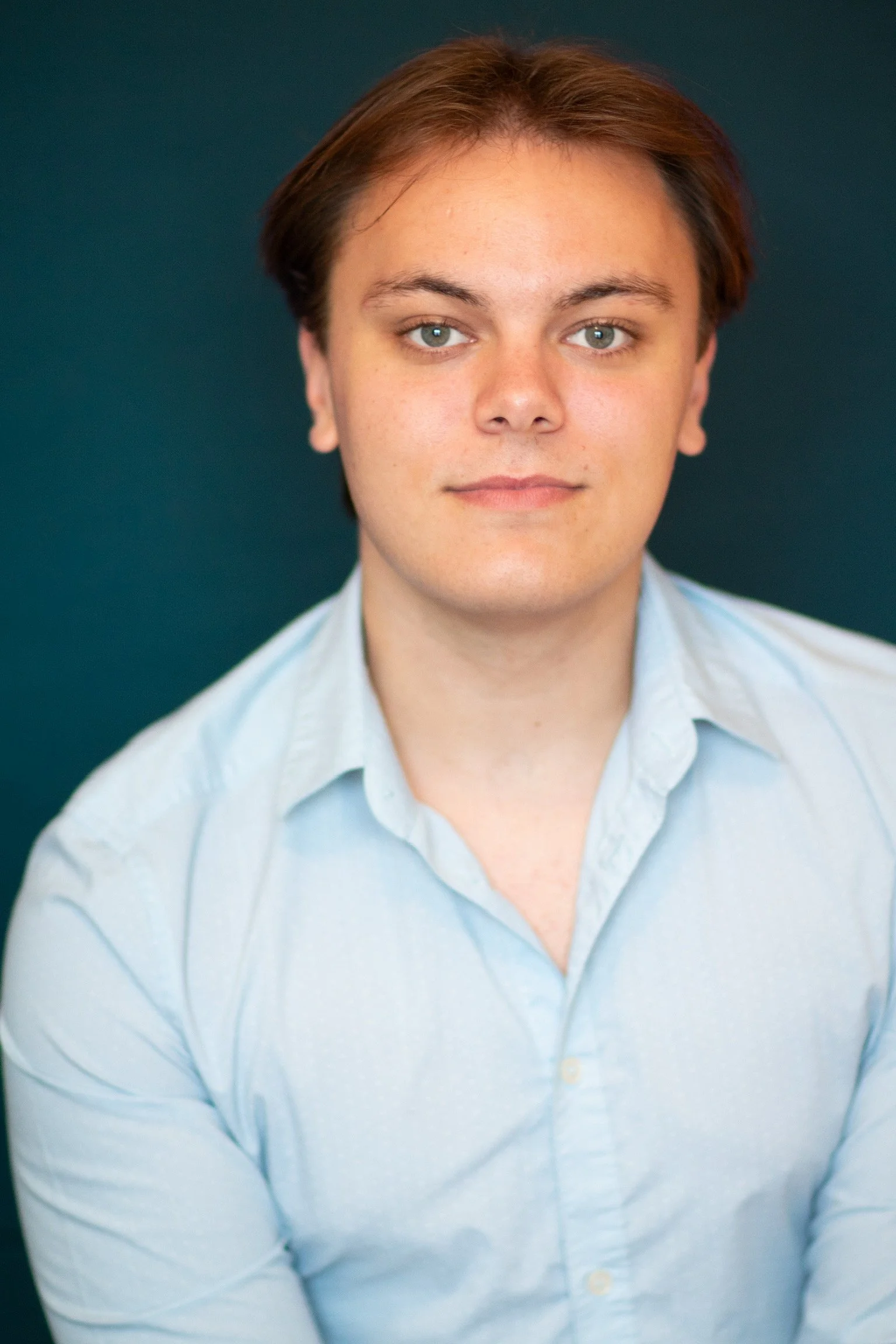 Portrait of a young man with light skin and brown hair, wearing a light blue collared shirt, against a dark teal background.