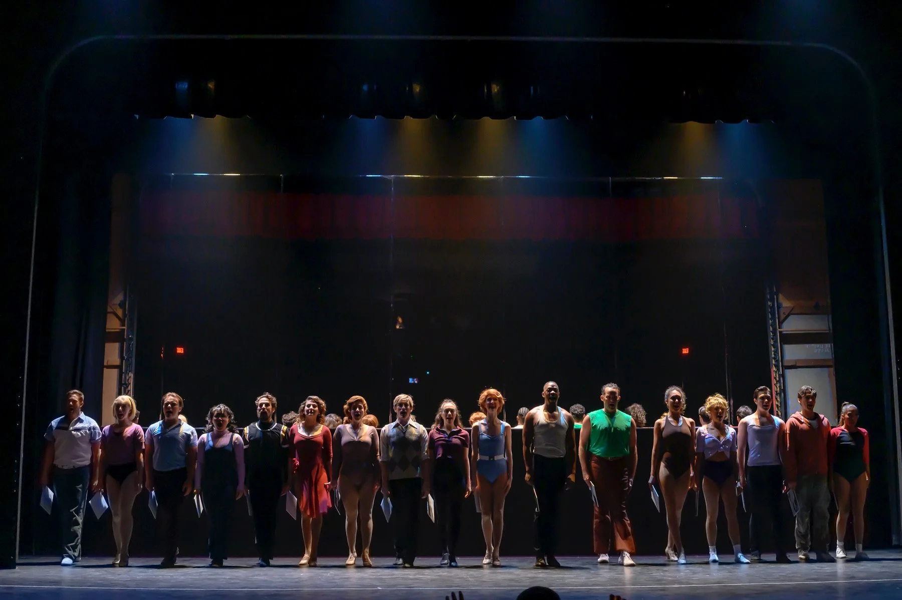 Stage with 16 performers standing in a line, lit by blue and yellow stage lights, with a dark background.