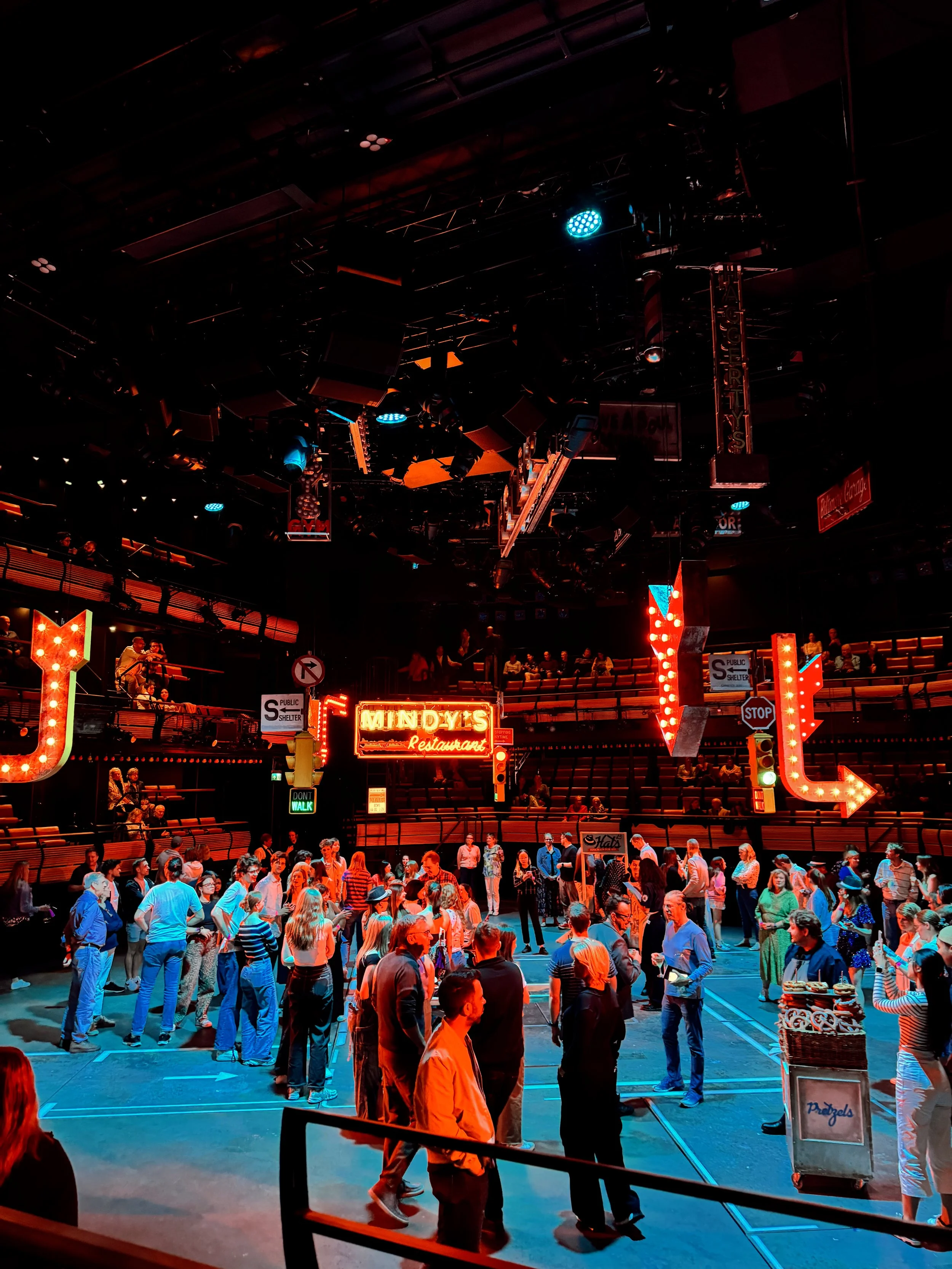 People gather inside a theater with neon signs and hanging overhead lights, with a large crowd standing and talking on the main floor and audience seating visible in the background.