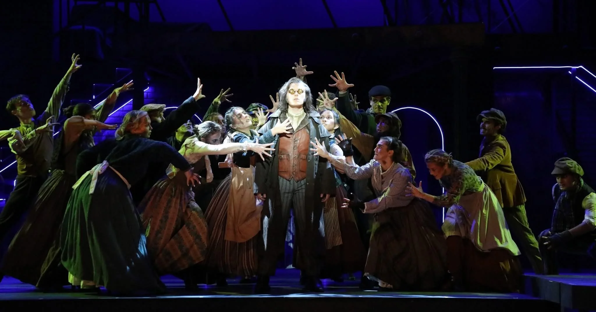 Theater scene with a woman wearing glasses at center, surrounded by a group of people in vintage clothing reaching toward her on stage with dramatic lighting and dark background.