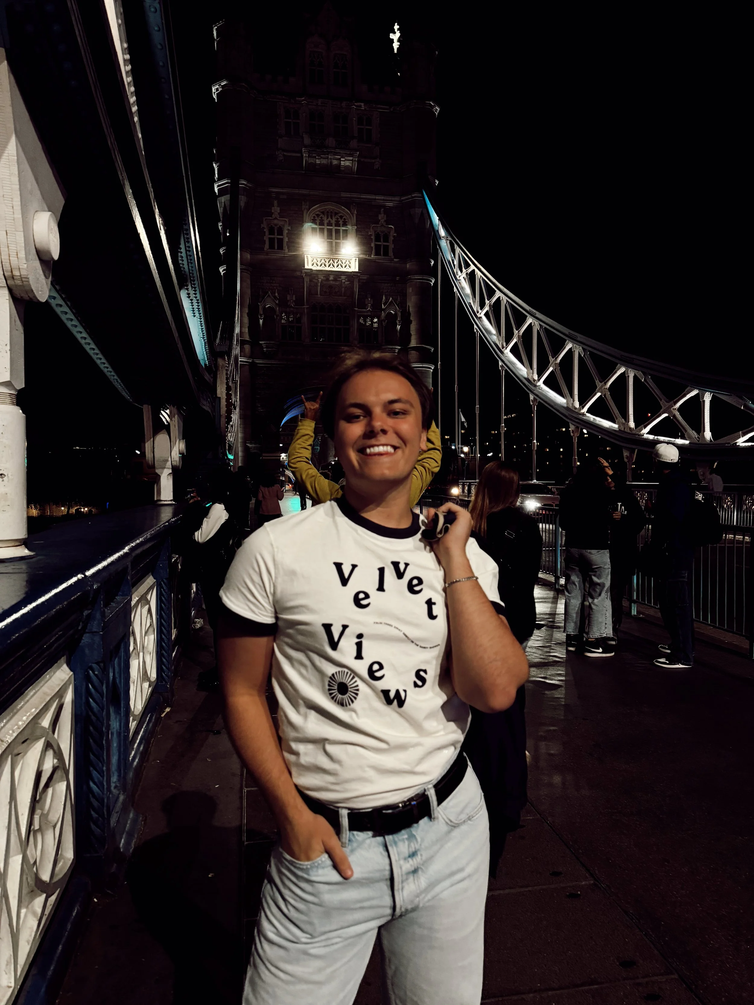 A smiling young person standing on a bridge at night, with the Tower Bridge illuminated in the background.