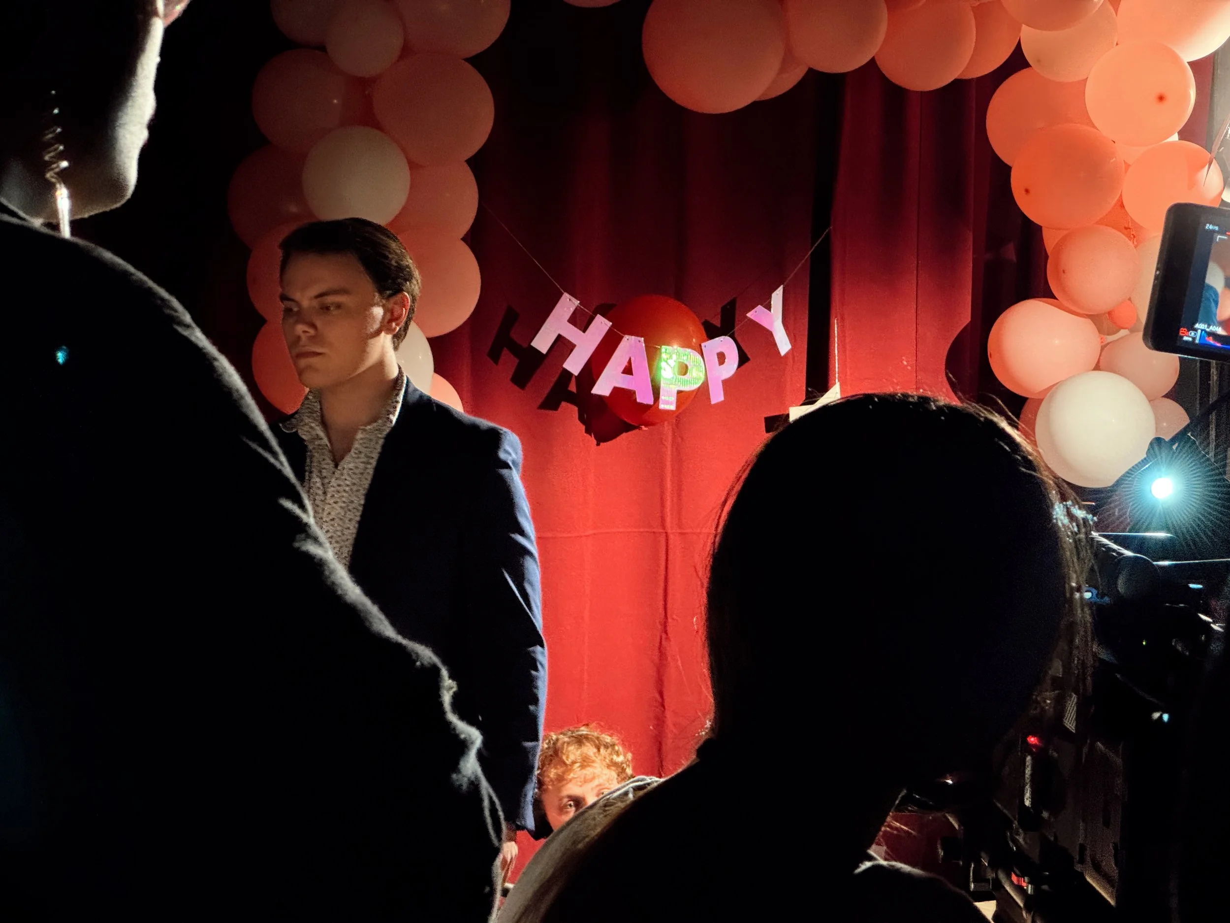 A shot from a film set with Christian at a celebration event with pink and white balloons and a 'HAPPY' banner, with a red backdrop and stage lighting.