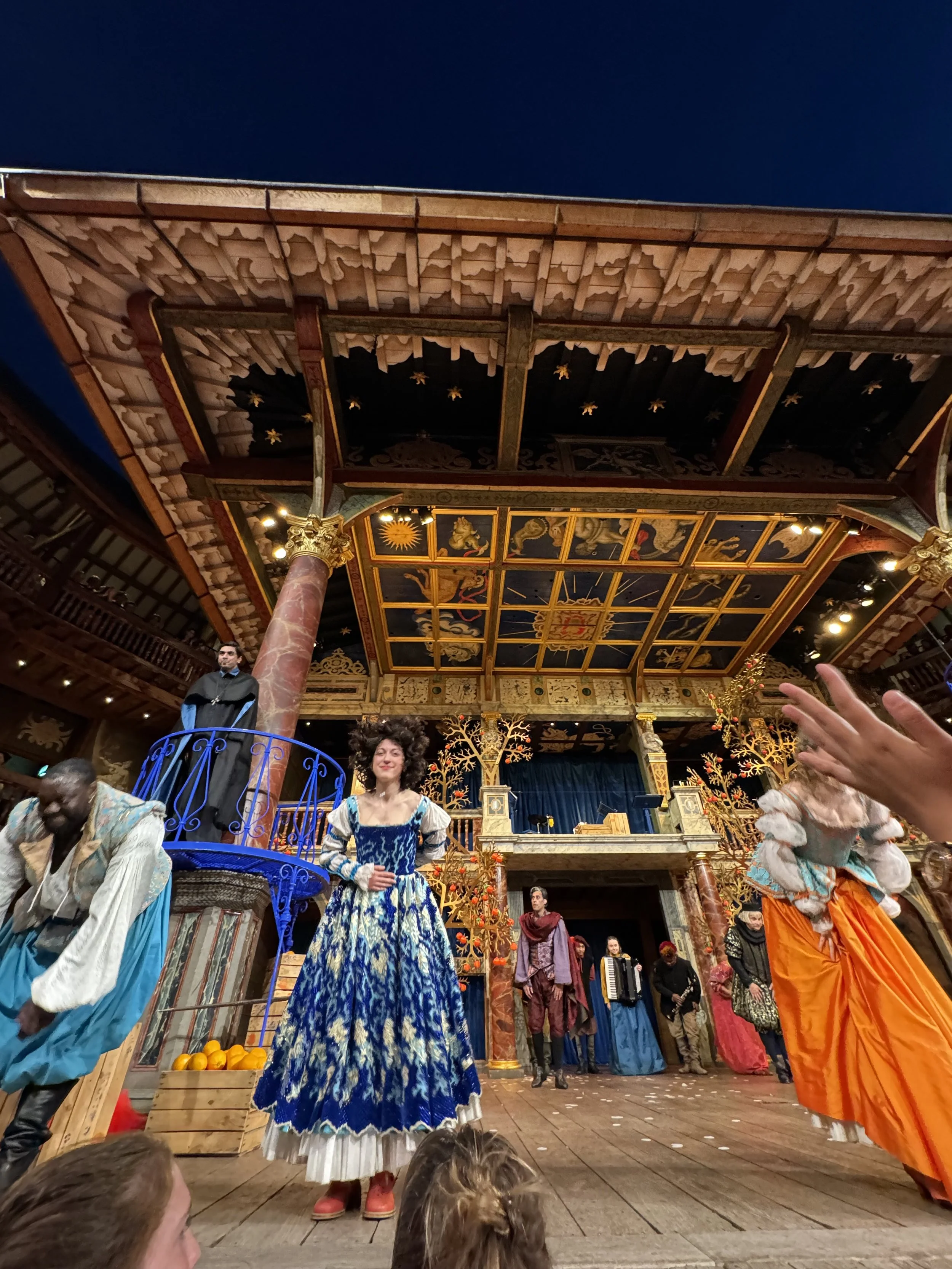 Scene from a theatrical play with actors dressed in colorful period costumes on a detailed stage with ornate decorations and painted ceiling.