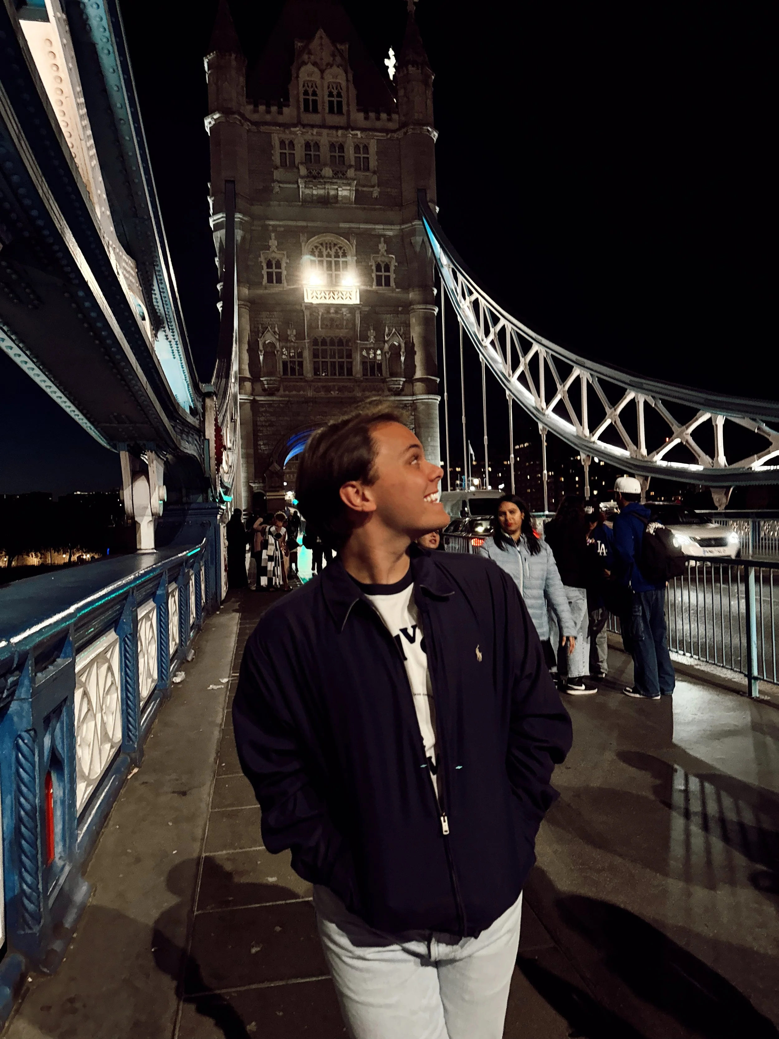 A young man standing on Tower Bridge in London at night, smiling and looking upwards, with the illuminated bridge and tower in the background.