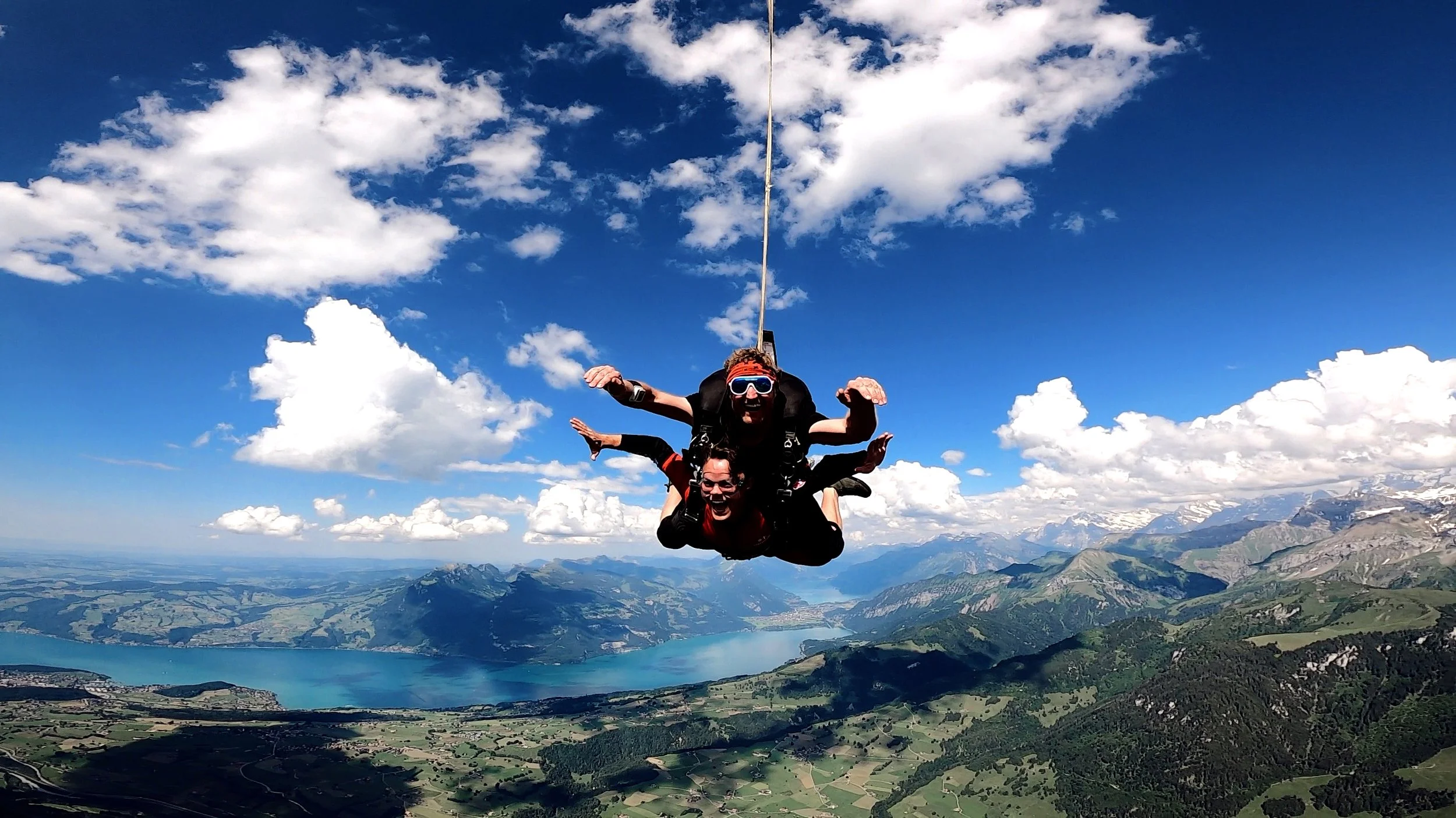 Two people skydiving in freefall over a mountainous landscape with a lake, under a partly cloudy sky.