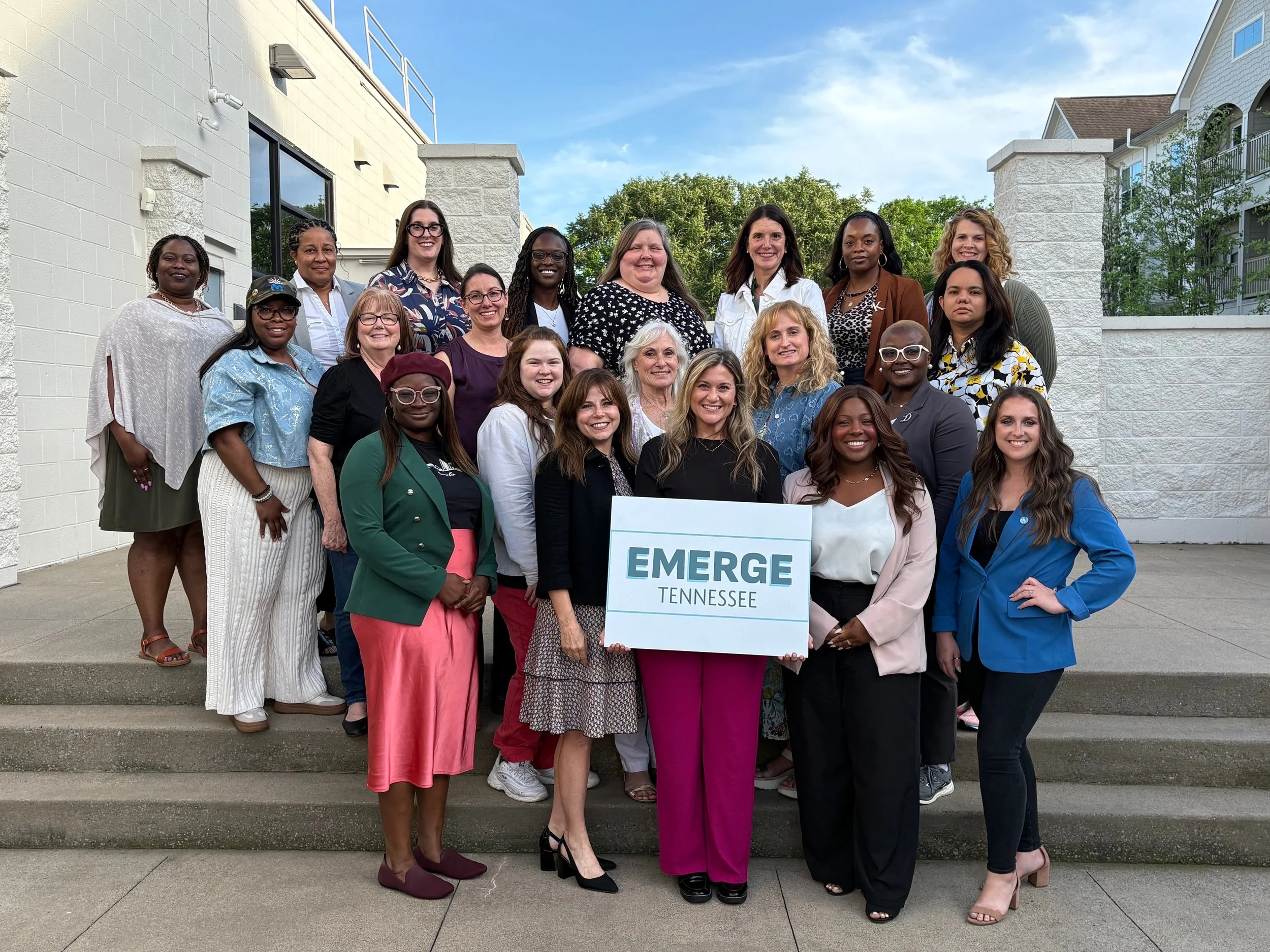 Group of diverse women standing on steps outside, holding a sign that reads 'EMERGE Tennessee' on a bright, sunny day.