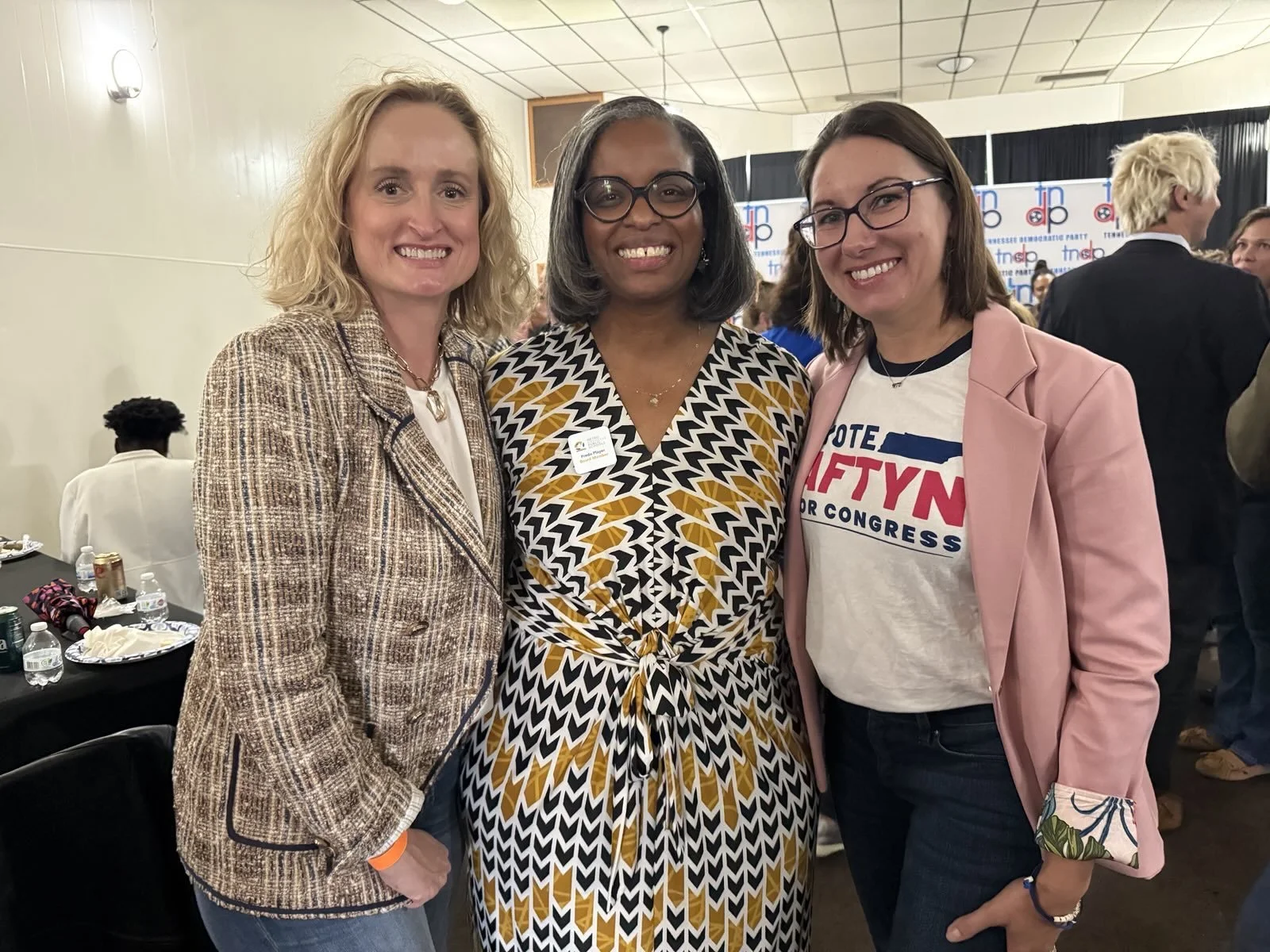 Three women standing together at a political event, smiling for the camera. The woman on the right is wearing a light pink blazer and a T-shirt with a political slogan. The woman in the middle is wearing a patterned dress, and the woman on the left is wearing a tweed jacket.