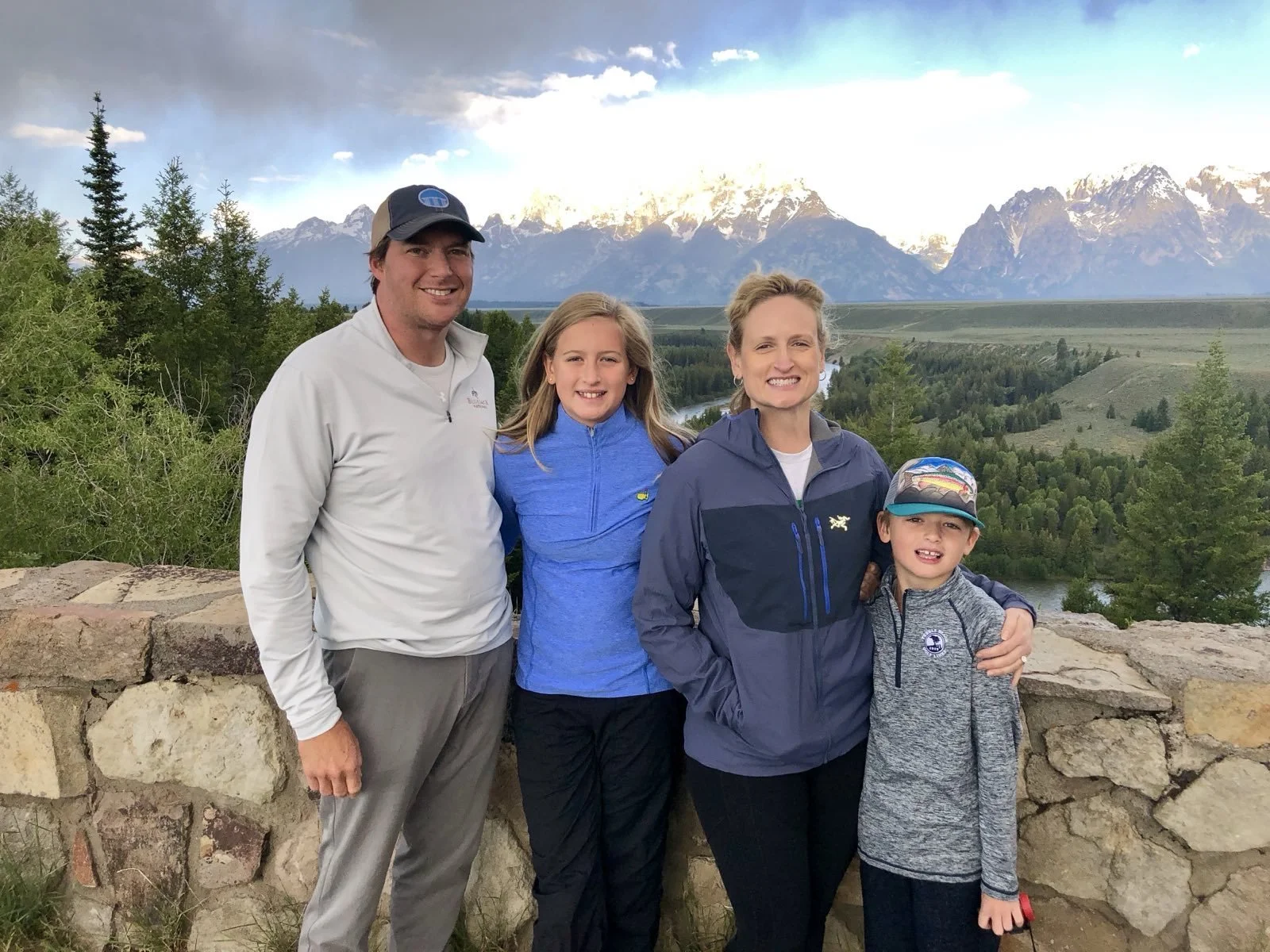 Family of four posing outdoors with mountains, trees, and a river in the background.