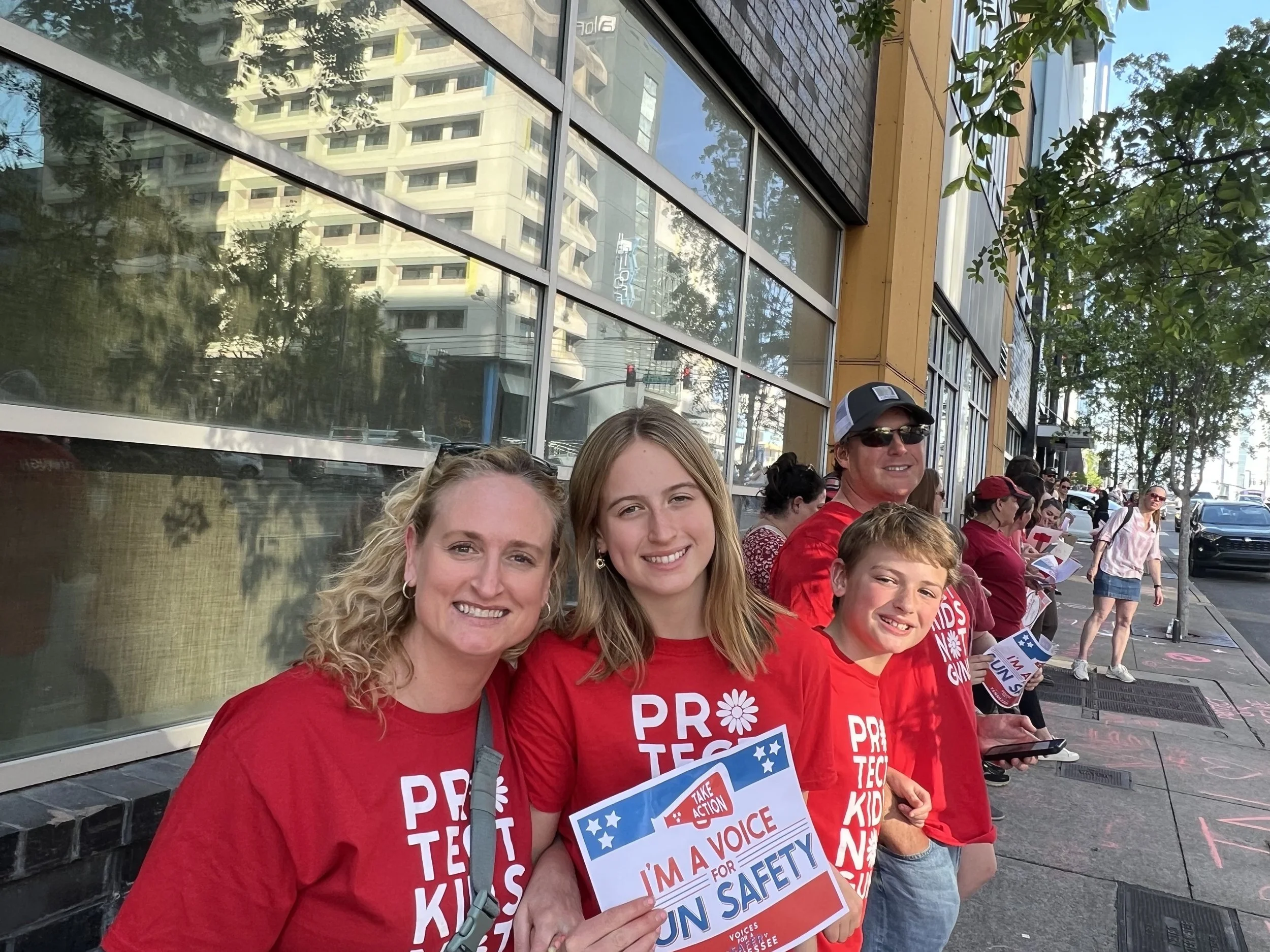 A group of people wearing red shirts with white text protesting or rallying on a city sidewalk. The woman in the front holds a sign that reads 'I'm a voice for gun safety.' The group includes children and adults, some wearing sunglasses and hats. The background shows a large glass window reflecting buildings and trees, with other protesters and pedestrians along the street.