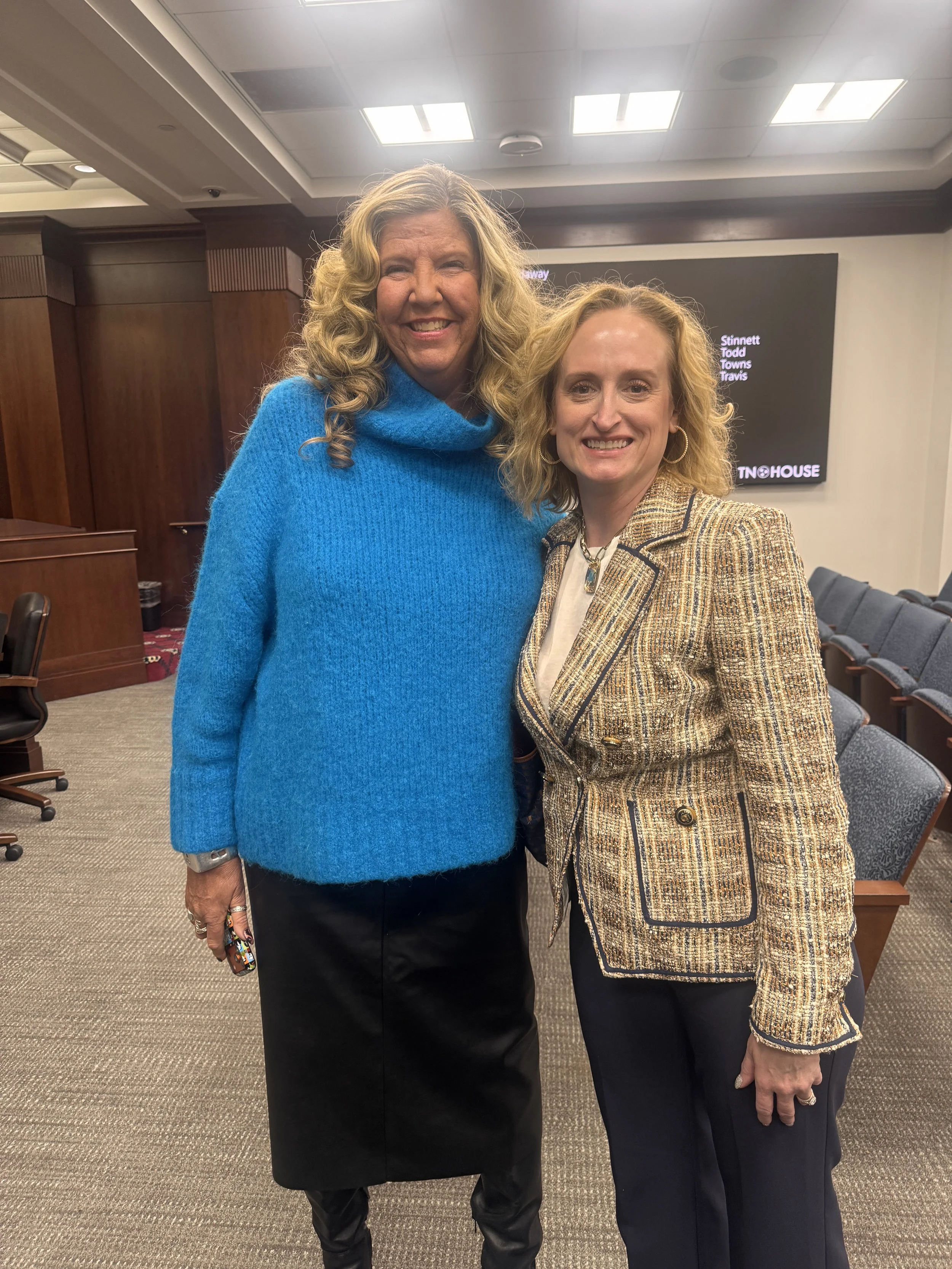 Two women standing in a room with wood-paneled walls, smiling at the camera. One woman is wearing a bright blue turtleneck sweater and a black skirt, while the other is in a patterned blazer and dark pants. There are rows of chairs and a large screen in the background.