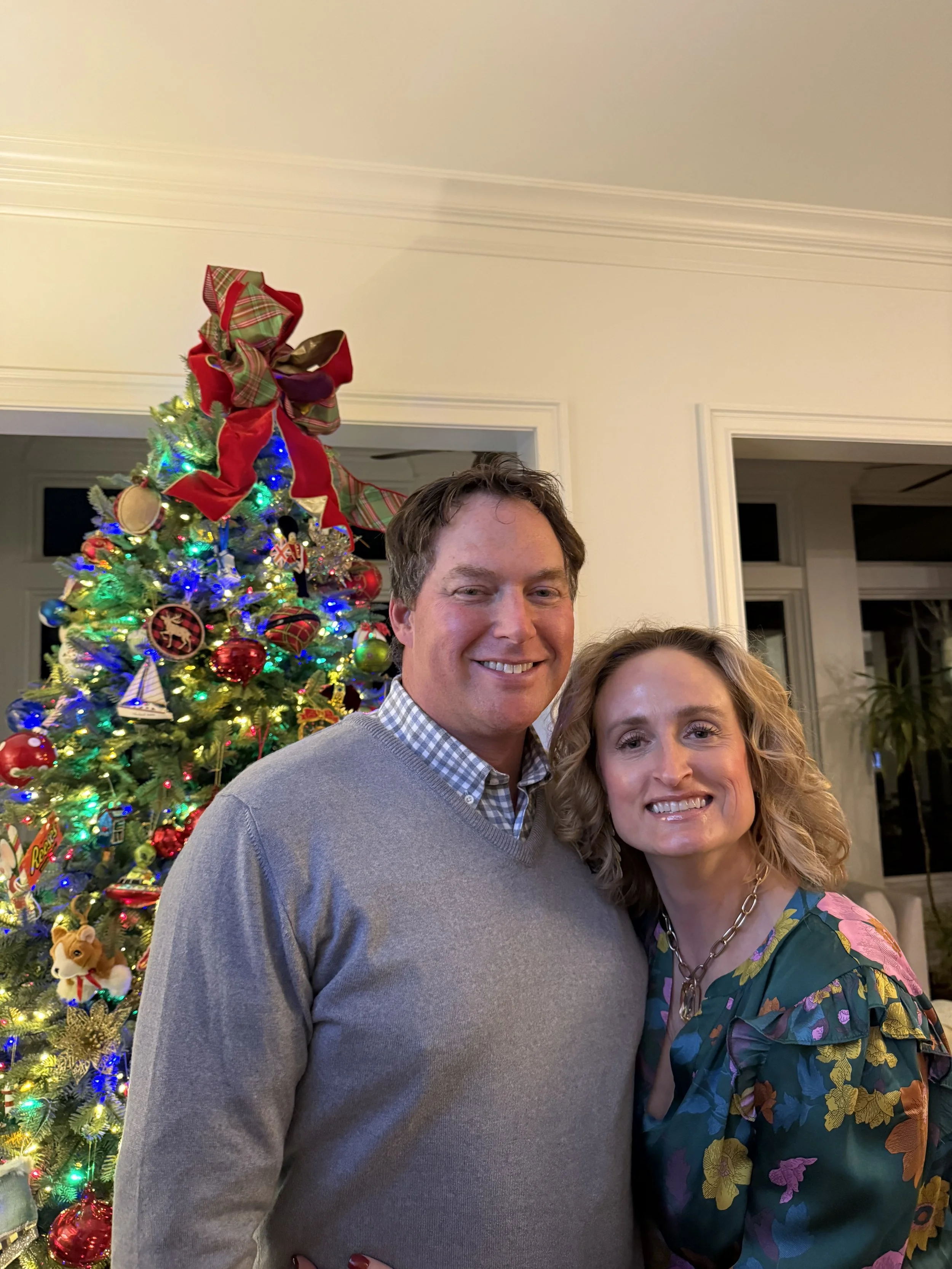 A smiling man and woman stand close together in front of a decorated Christmas tree with colorful lights and ornaments.