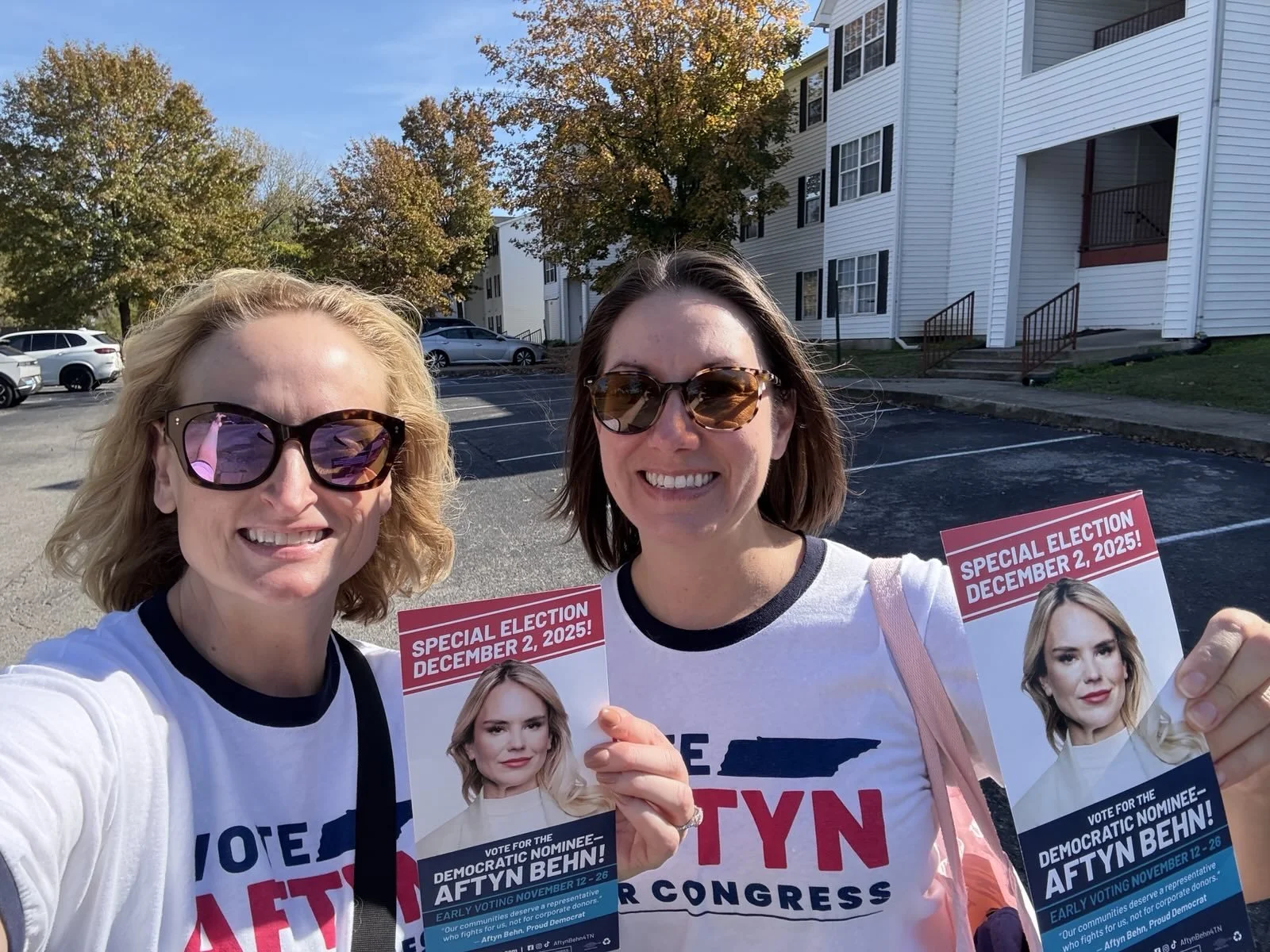 Two women standing outside in a parking lot holding campaign flyers for the Democratic nominee, AFTYN BEHN, for special election on December 2, 2025. Both women are smiling and wearing sunglasses, with buildings and trees in the background.