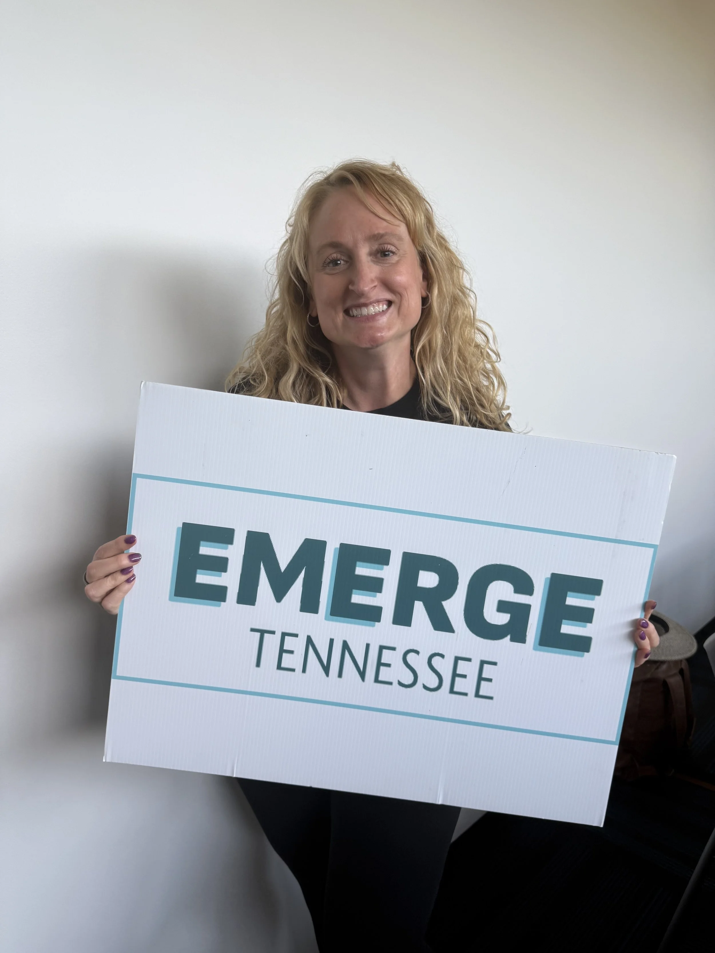 A woman with curly blonde hair smiling and holding a sign that says 'EMERGE TENNESSEE' in large letters.
