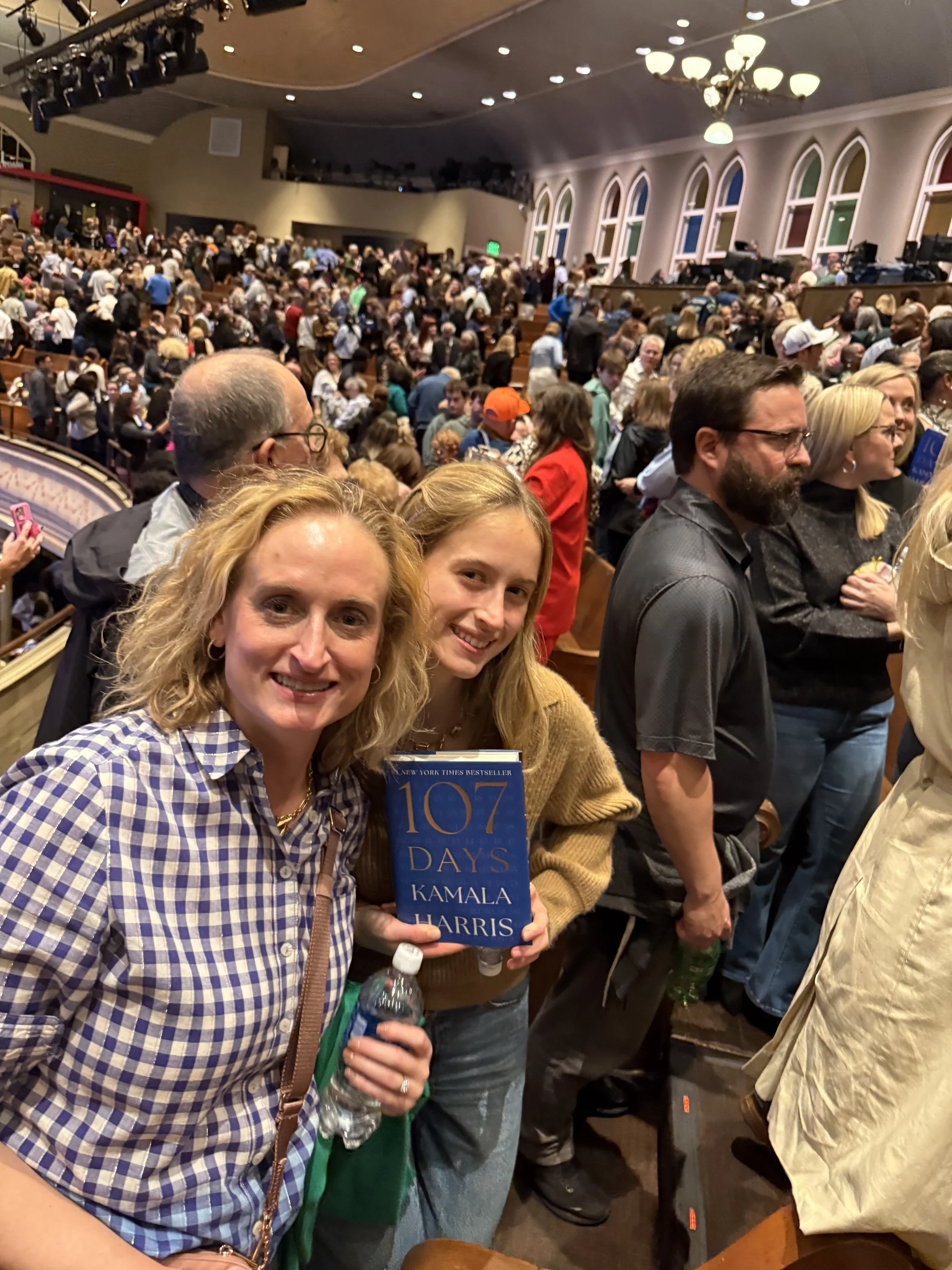 A large crowd in an auditorium, with two women in the foreground holding a book titled '107 Days' by Kamala Harris, smiling for the camera.