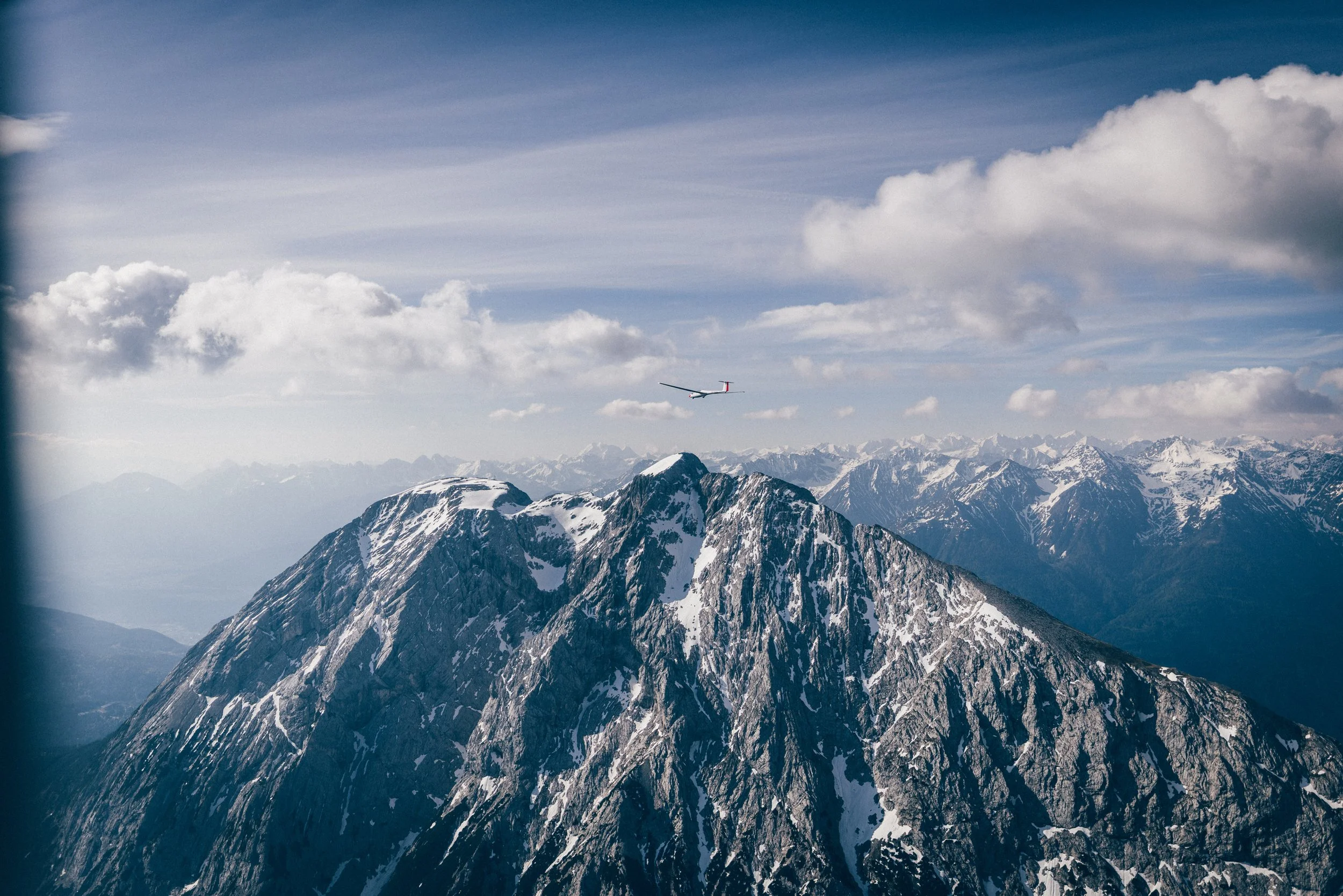 Berggipfel mit Schneefeldern, Flugzeug in der Luft, Wolken am Himmel, im Hintergrund weitere Bergketten.