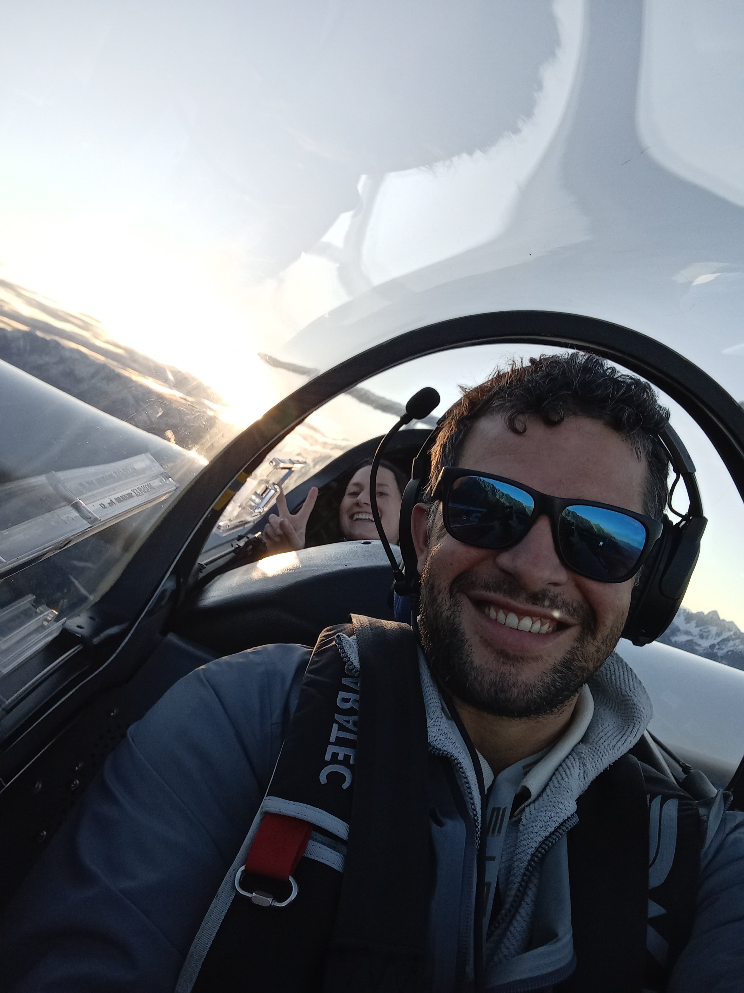 Zwei Personen in einem Flugzeug, der Mann vorne trägt Sonnenbrille und Kopfhörer, die Frau im Hintergrund macht Peace-Zeichen. Sonnenuntergang oder Sonnenaufgang im Hintergrund.