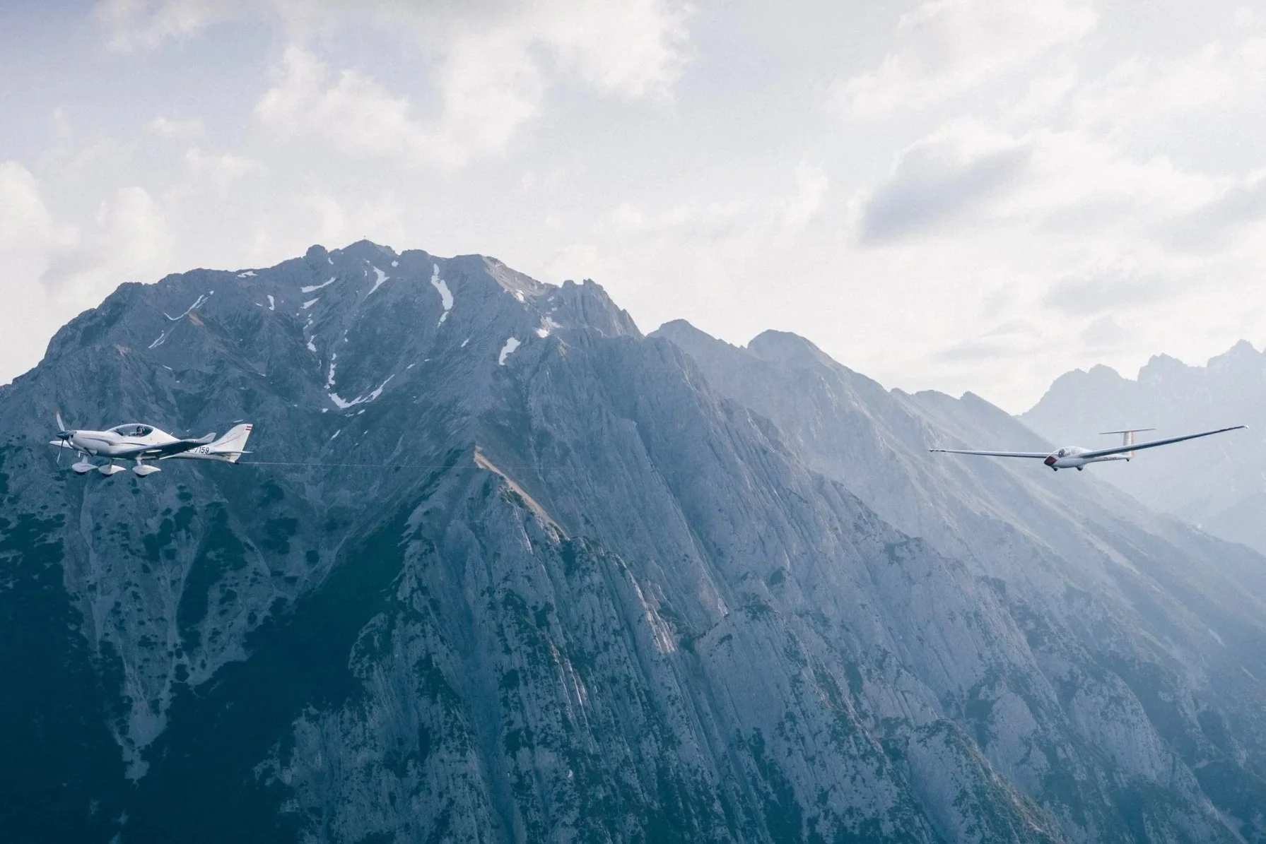 Ein Flugzeug und ein segelflugzeug fliegen vor einer Bergkulisse mit hohen, teilweise schneebedeckten Bergen in den Wolken.