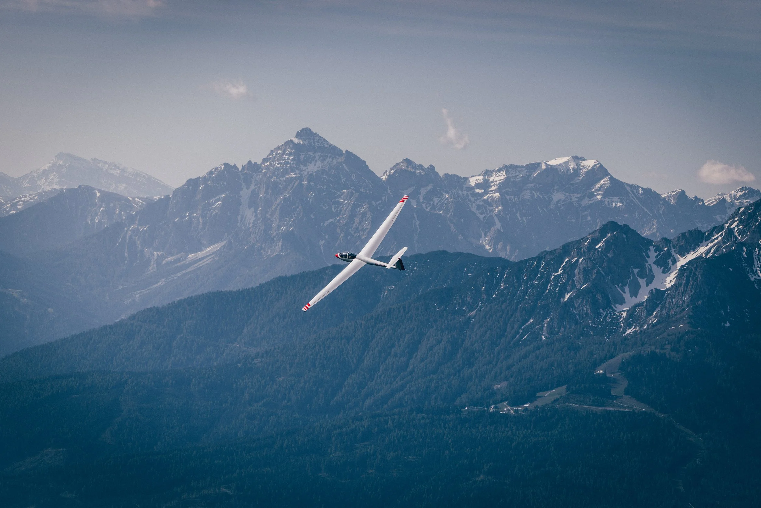 Segelflugzeug fliegt über die Berge mit Schnee auf den Gipfeln und bewaldeten Hängen.