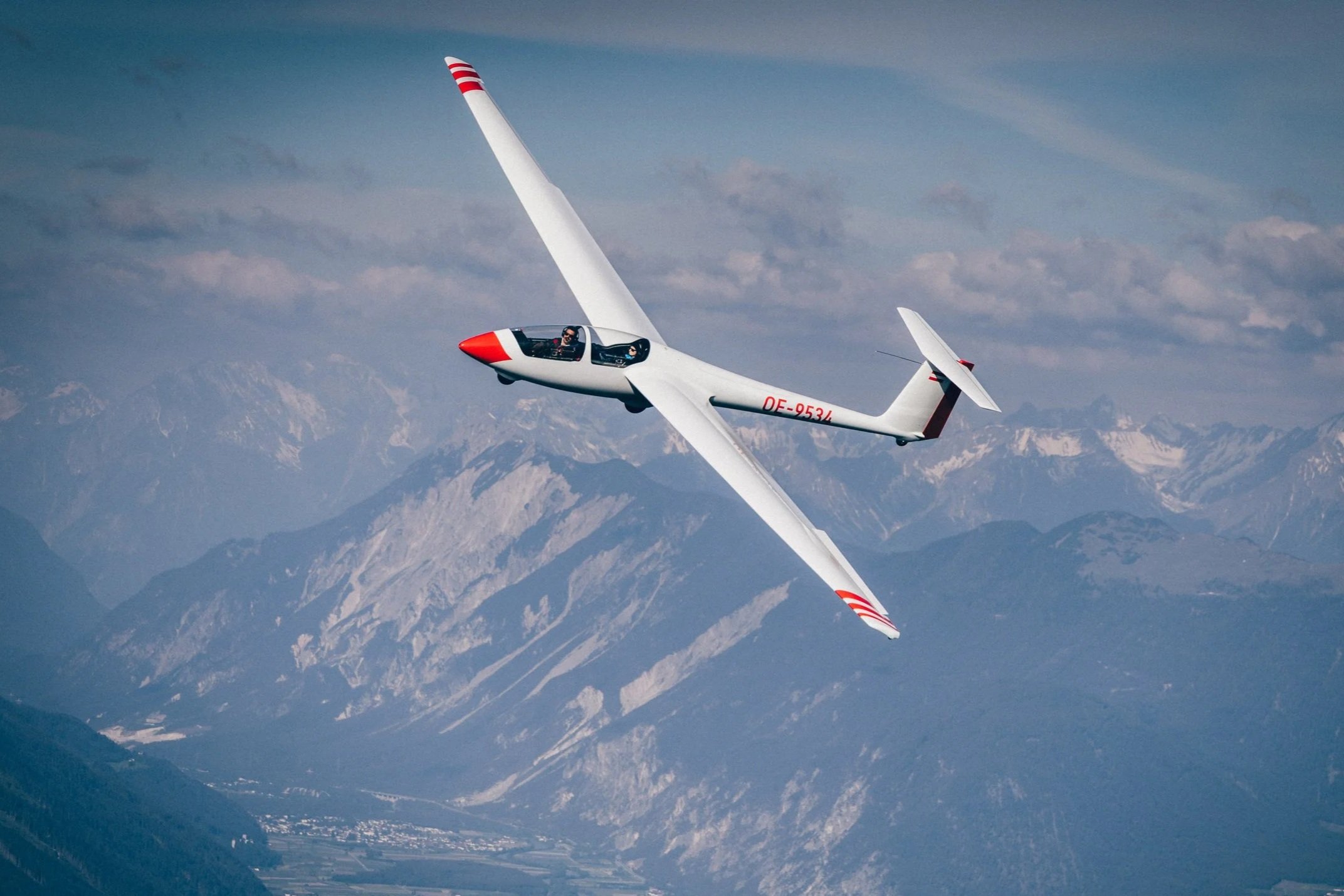 Ein weißes Segelflugzeug mit rotem Schnabel fliegt über eine Berglandschaft mit schneebedeckten Gipfeln.