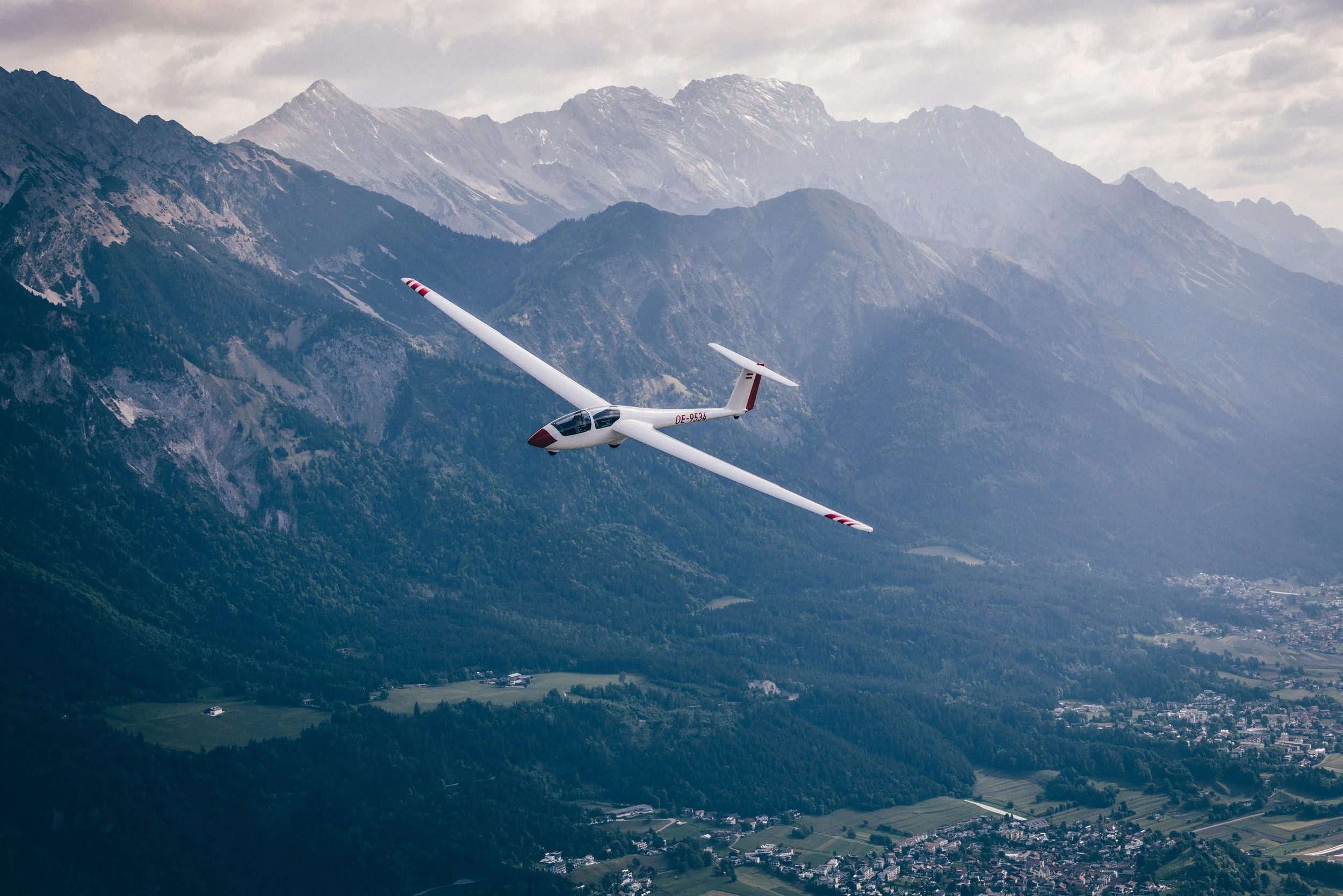Segelflugzeug fliegt über eine Berglandschaft mit Hügeln, Wäldern und Dörfern, umgeben von hohen Bergen.