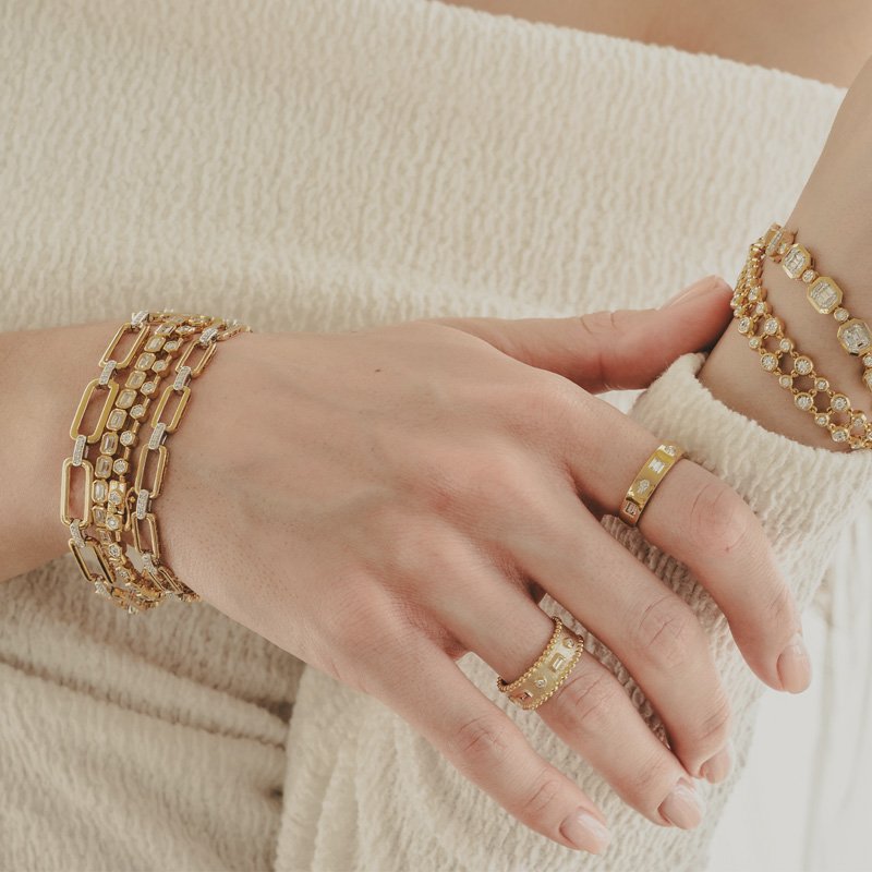 A person's hand and wrist adorned with multiple gold and diamond jewelry pieces, including bracelets and rings, resting on a cream-colored fabric.
