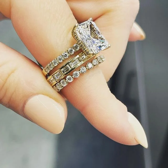 Close-up of a hand wearing three rings on the ring finger, with a large rectangular diamond engagement ring and two wedding bands, one with small diamonds and the other with rectangular embellishments.