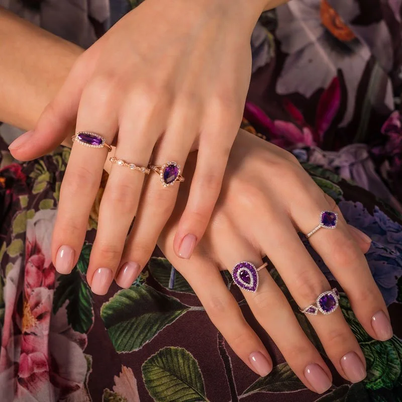 Two hands displaying multiple Fashion rings withcolorful gemstone and decorative designs, against a floral-patterned dress.