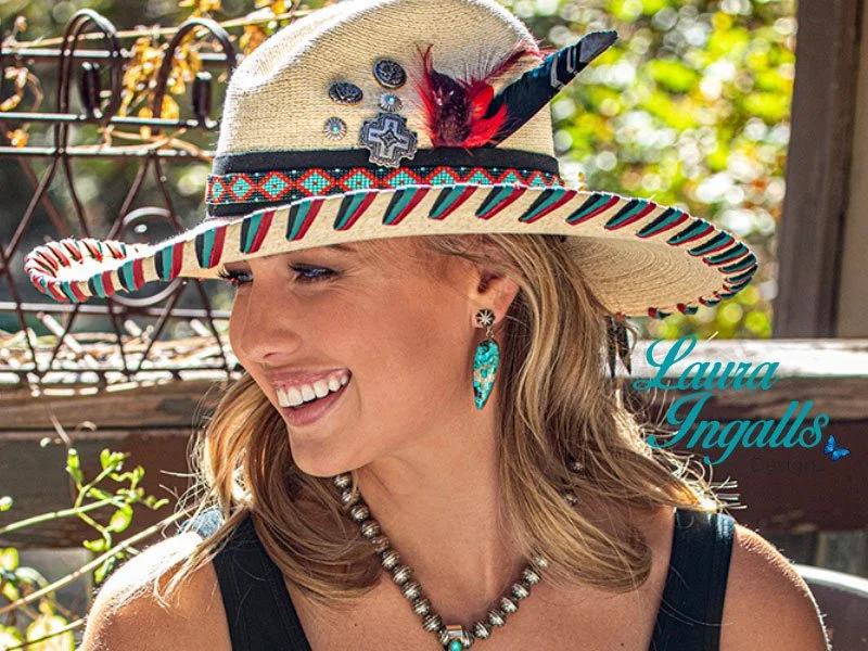 A woman smiling outdoors wearing a wide-brimmed hat with decorations, turquoise earrings, a pearl necklace, and a black top.
