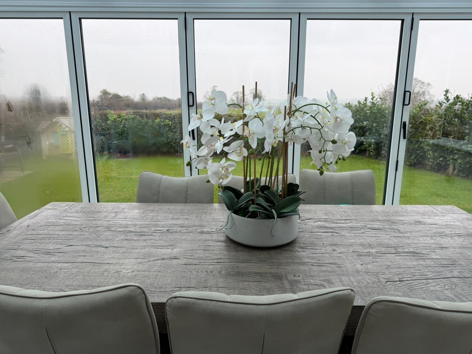 A dining table with a white orchid plant in a white pot, surrounded by beige chairs, situated in a glass sunroom with a view of a rainy backyard and cloudy sky.