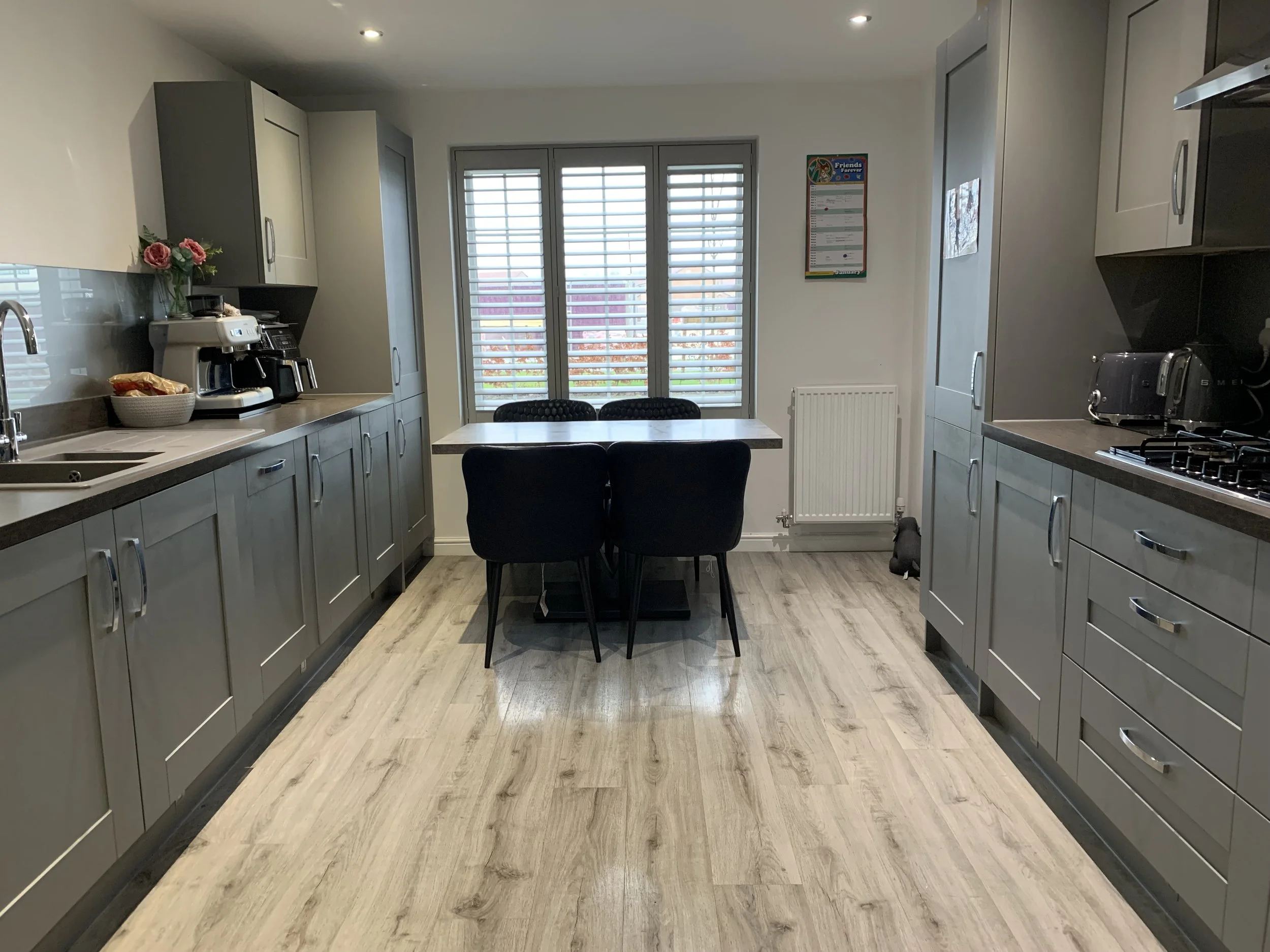 A modern kitchen with gray cabinets, a wooden floor, a dining table with four black chairs, a window with shutters, and kitchen appliances including a coffee machine, toaster, and stove.
