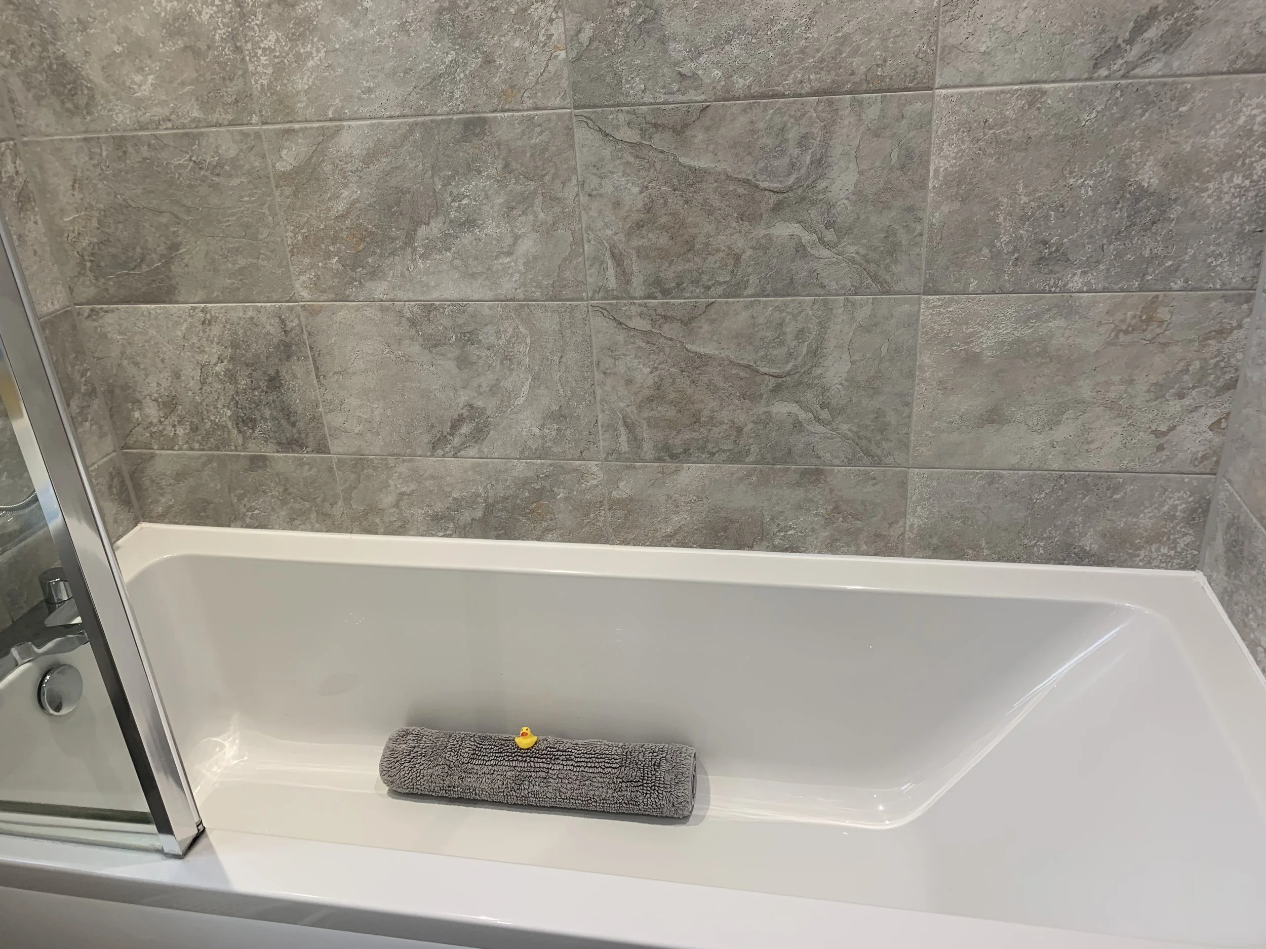 A white bathtub with a gray towel and a small rubber duck on top. The tub is surrounded by marbled gray tiled walls.