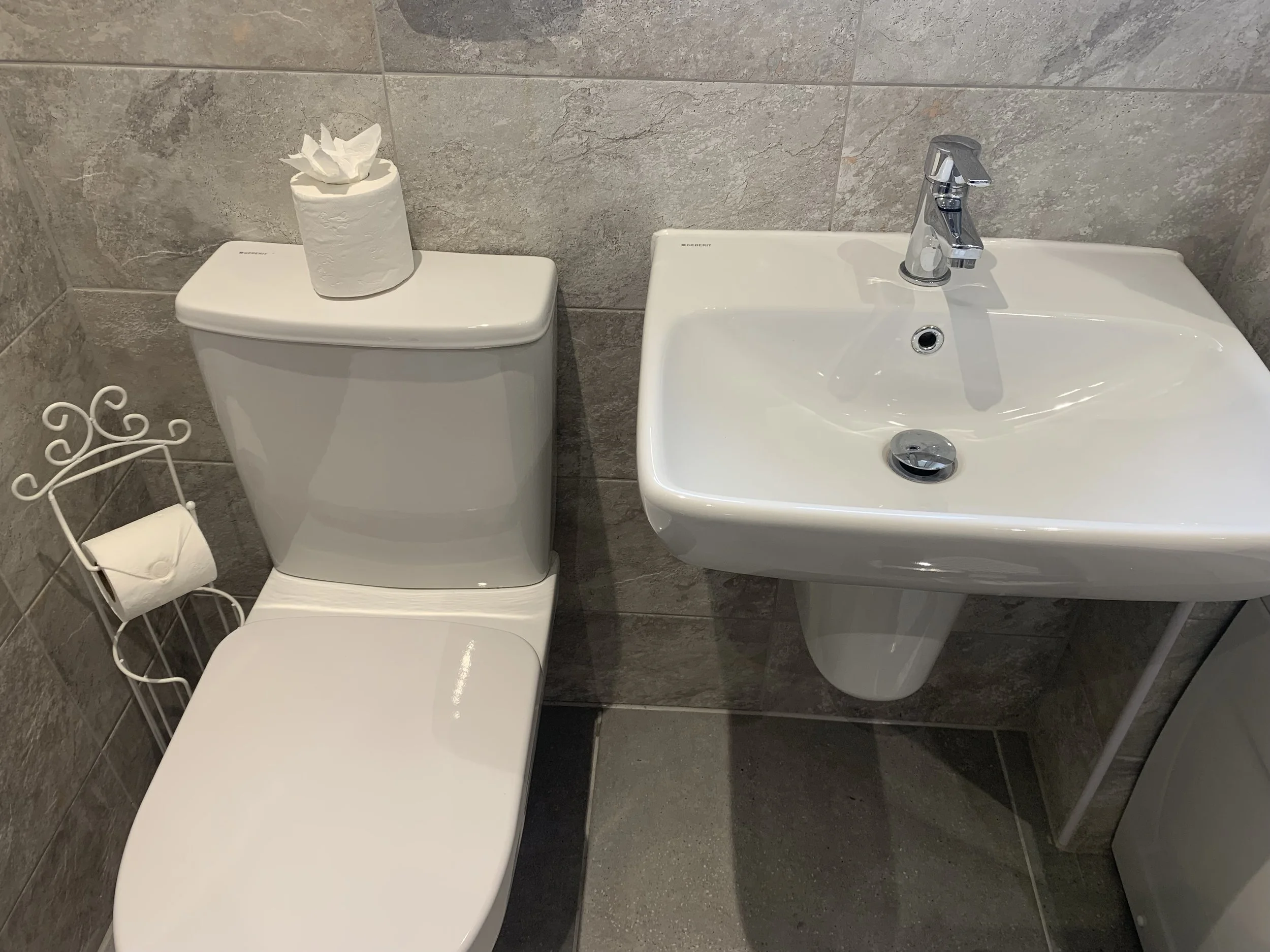A small bathroom with a white toilet, a white sink with a chrome faucet, a roll of toilet paper on a decorative metal holder, and a tissue box on top of the toilet tank. The bathroom has gray tiled walls and a gray tiled floor.