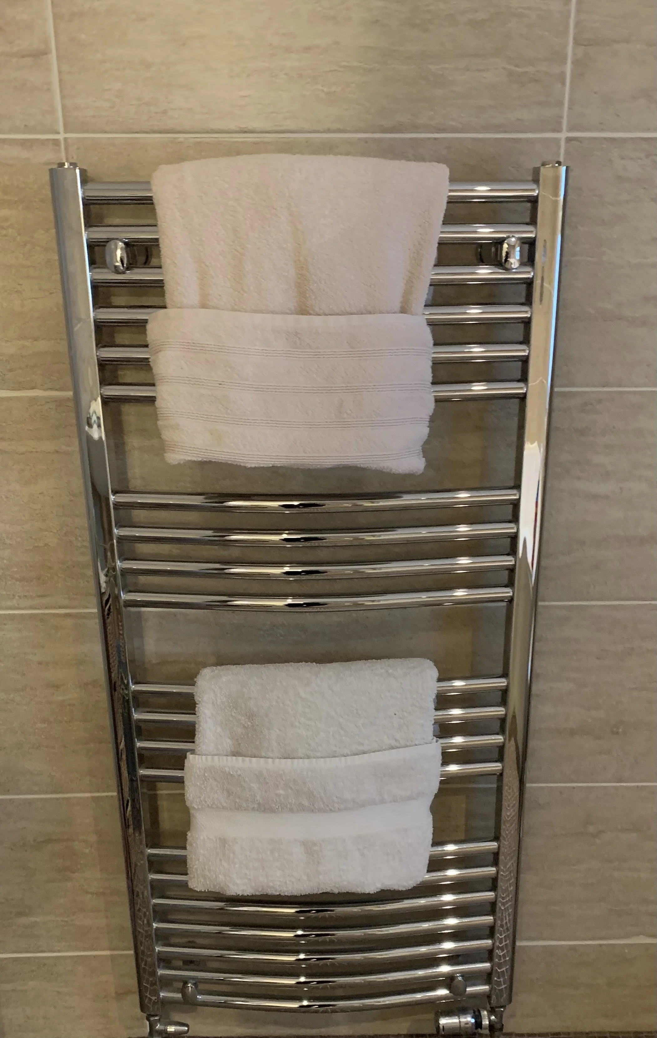 A stainless steel towel radiator with two neatly folded white towels hanging on it, mounted on a beige tiled wall.