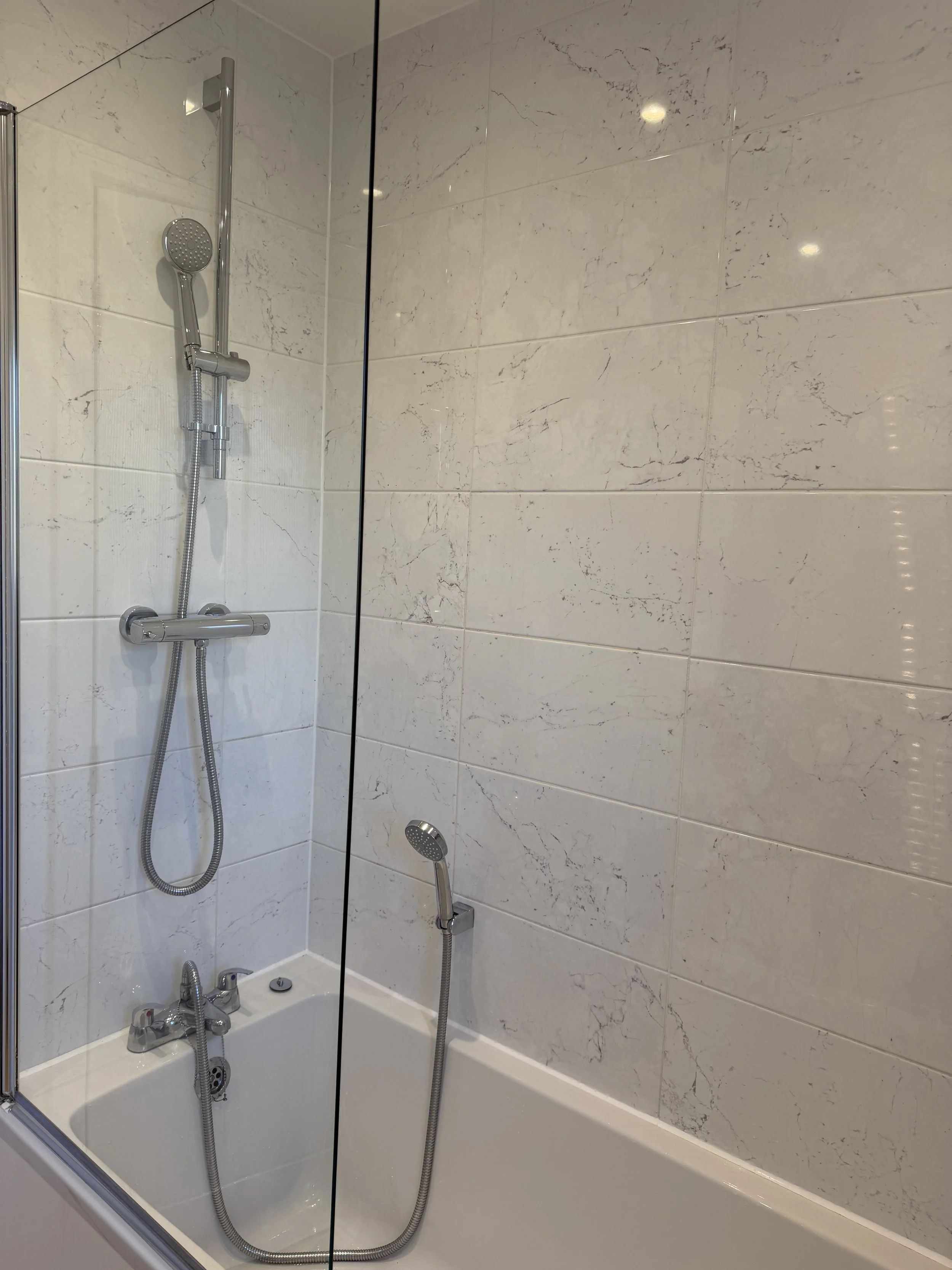A bathroom shower enclosure with a glass divider, a wall-mounted showerhead, a handheld shower, a bathtub, and white marble wall tiles.