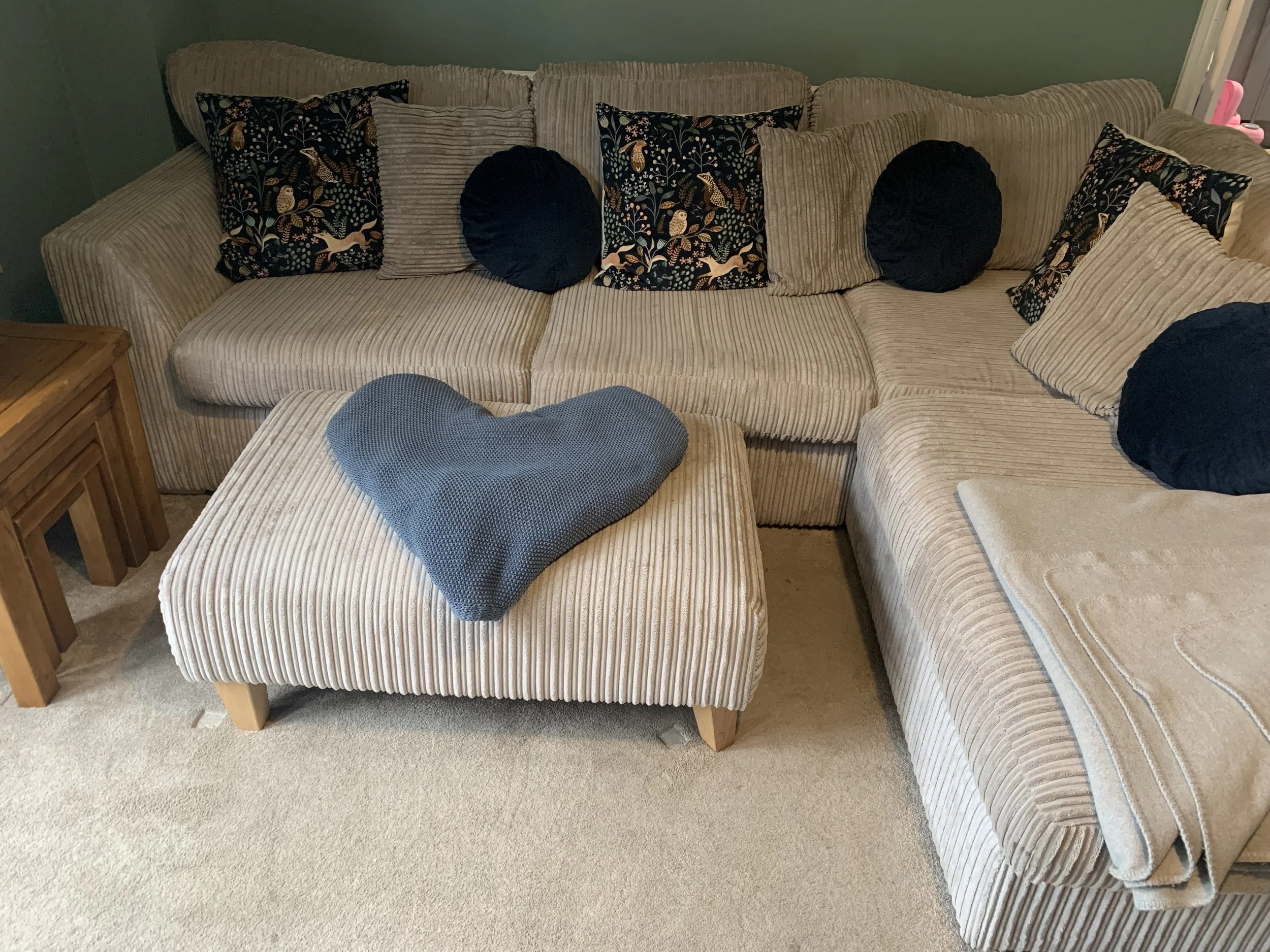 Living room with beige striped sectional sofa, decorated with black round and floral pillows, a gray heart-shaped cushion on an ottoman, and a beige blanket on the armrest. Small wooden side table on the left.