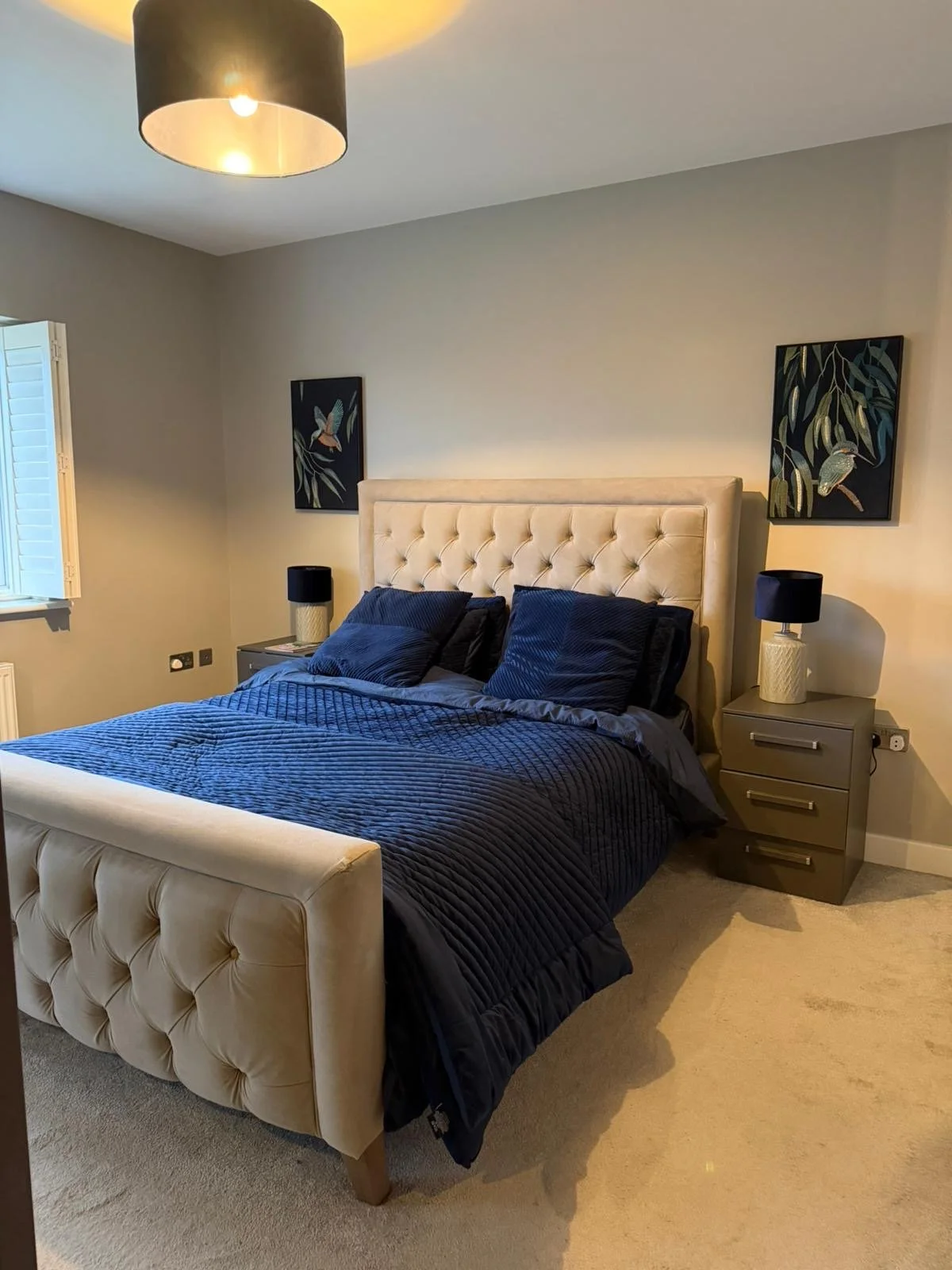 A bedroom with a beige tufted headboard, dark blue bedding, and two matching navy pillows. There are two gray nightstands with black lamps on each side of the bed. The room has light-colored walls with two framed bird and leaf artwork above each nigh