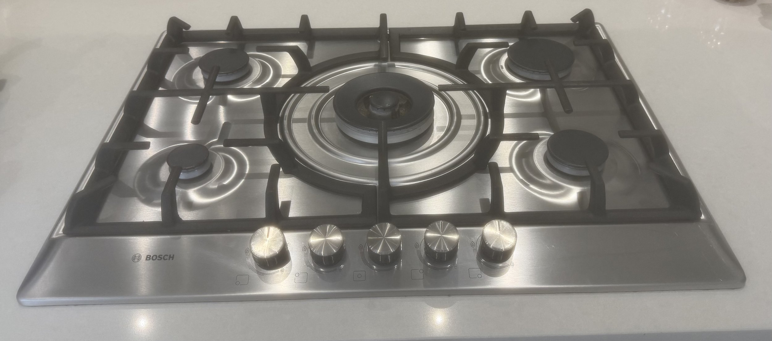 Stainless steel gas cooktop with five burners and control knobs in front, branded Bosch, on a white countertop.